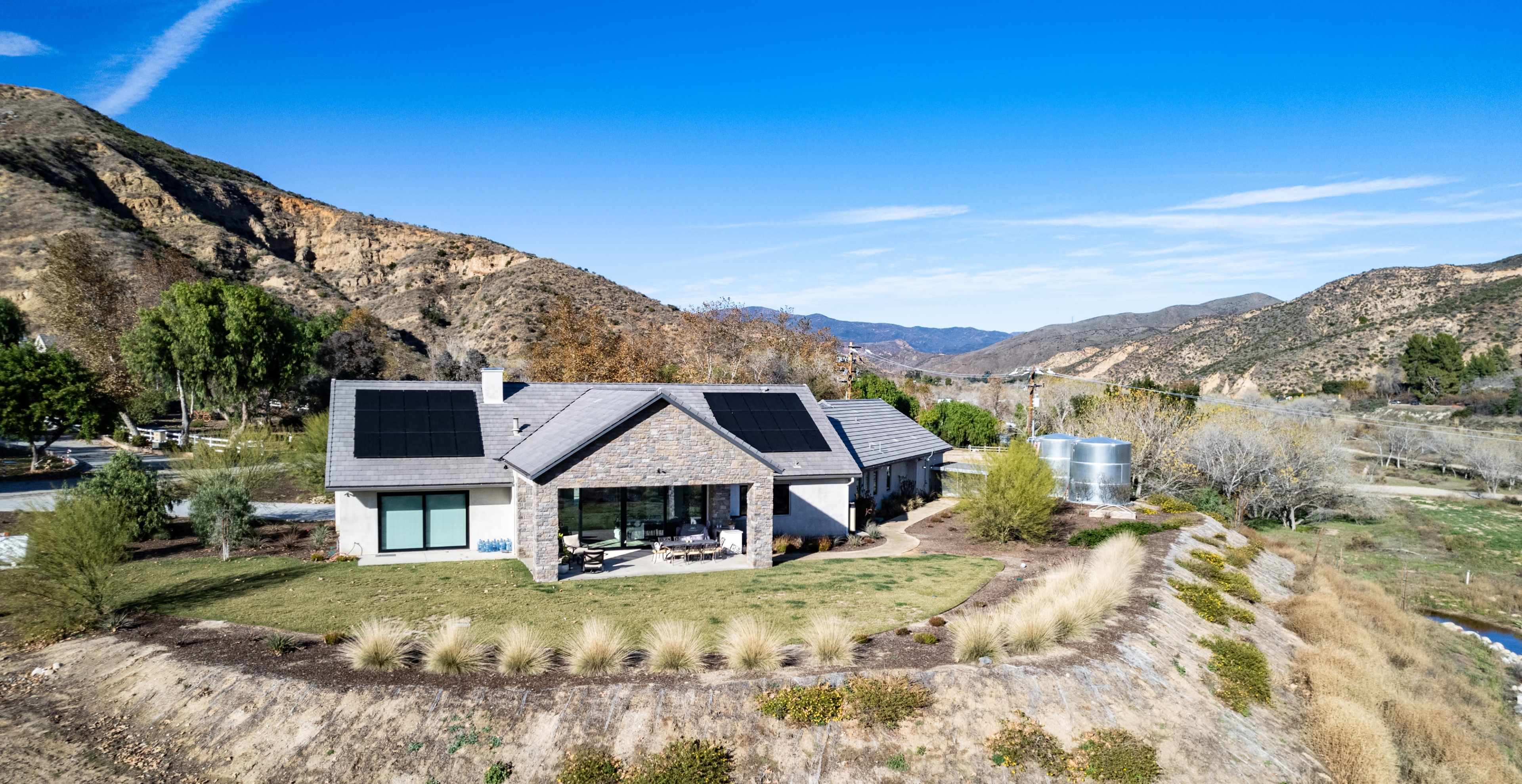 A modern house with solar panels is situated on a hillside, surrounded by vegetation and mountains.