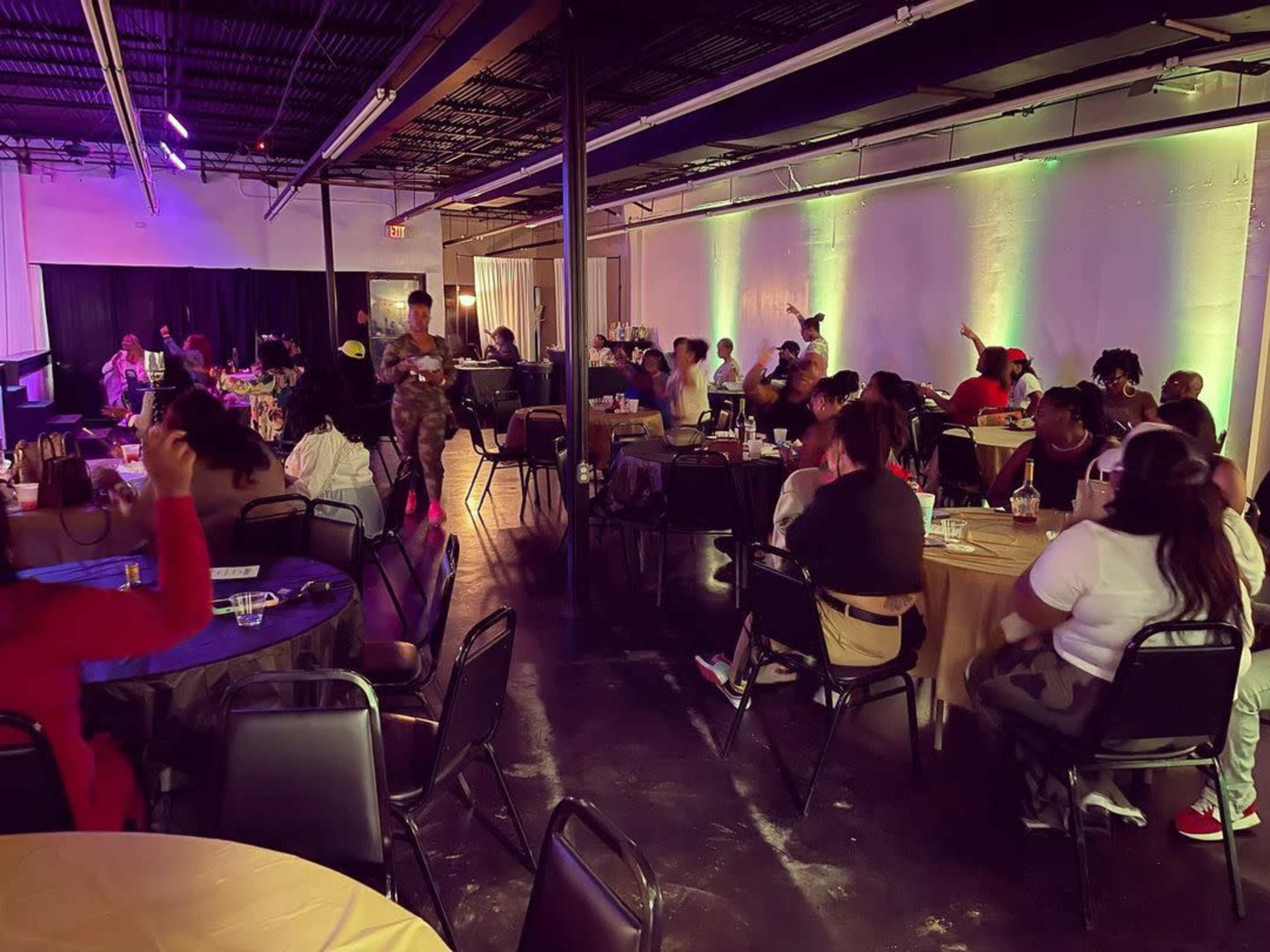 A lively indoor event with people seated at round tables, some engaging in conversation and others dancing in a well-lit space.