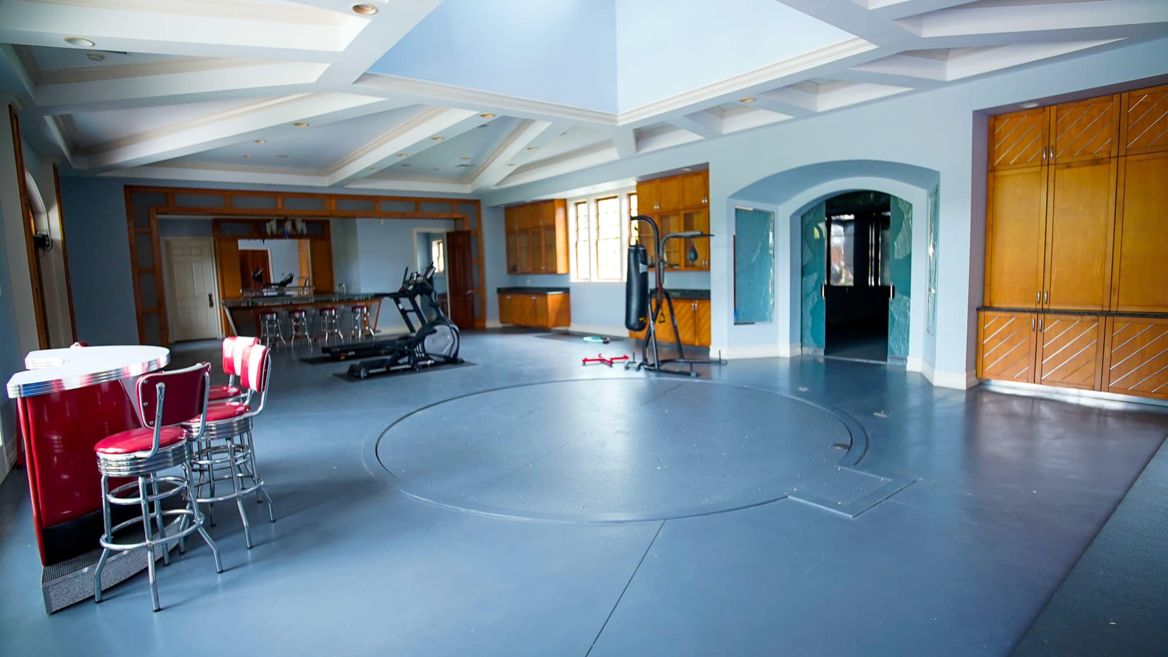 A spacious room with a gym area, exercise equipment, a bar with seating, and wooden cabinets along the walls.