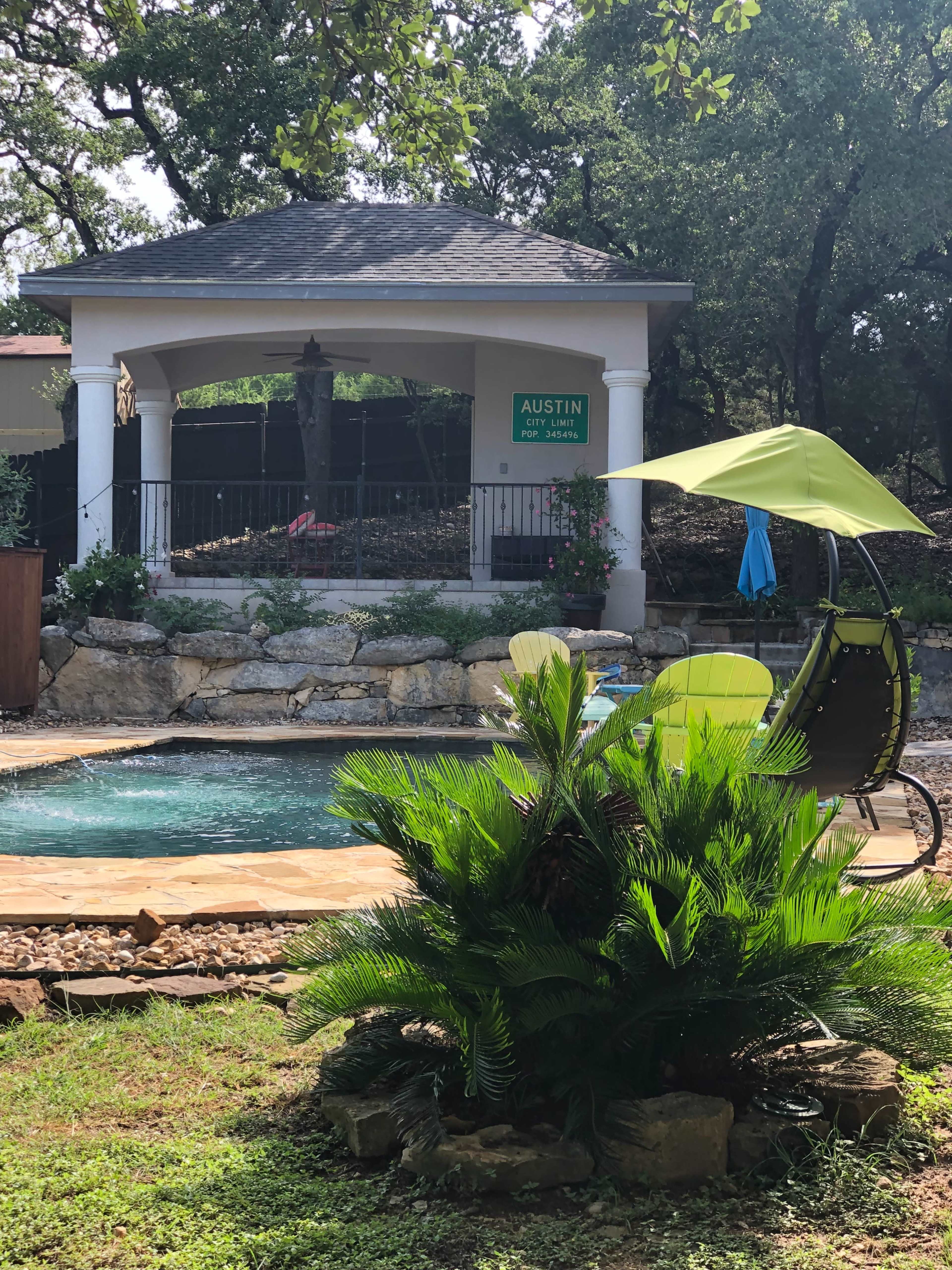 The scene features a circular pool surrounded by a stone deck, a green plant in the foreground, and a gazebo in the background with a sign that reads "AUSTIN."