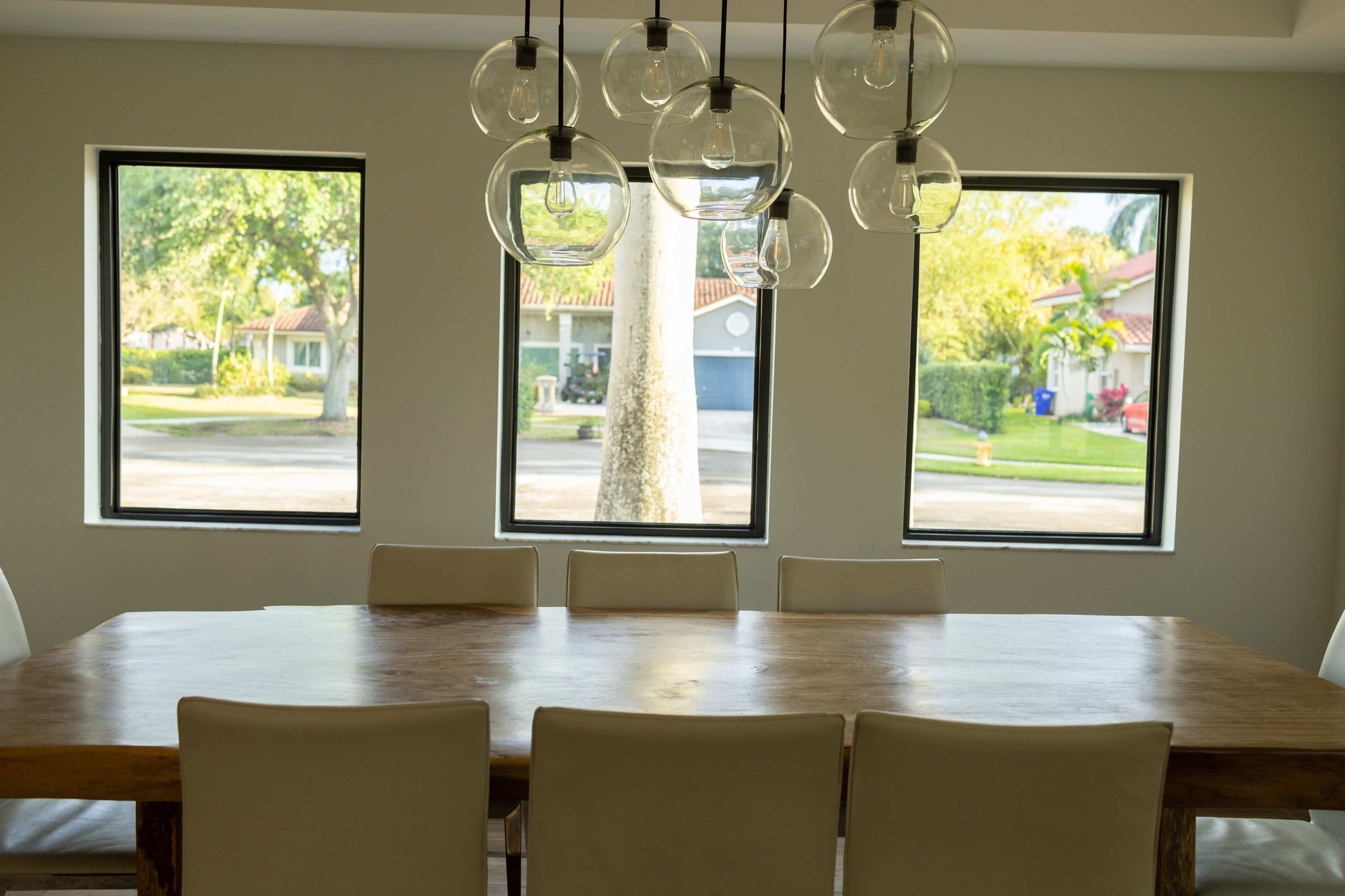 A wooden dining table with white chairs is illuminated by pendant lights, positioned near large windows that overlook a suburban street and greenery outside.