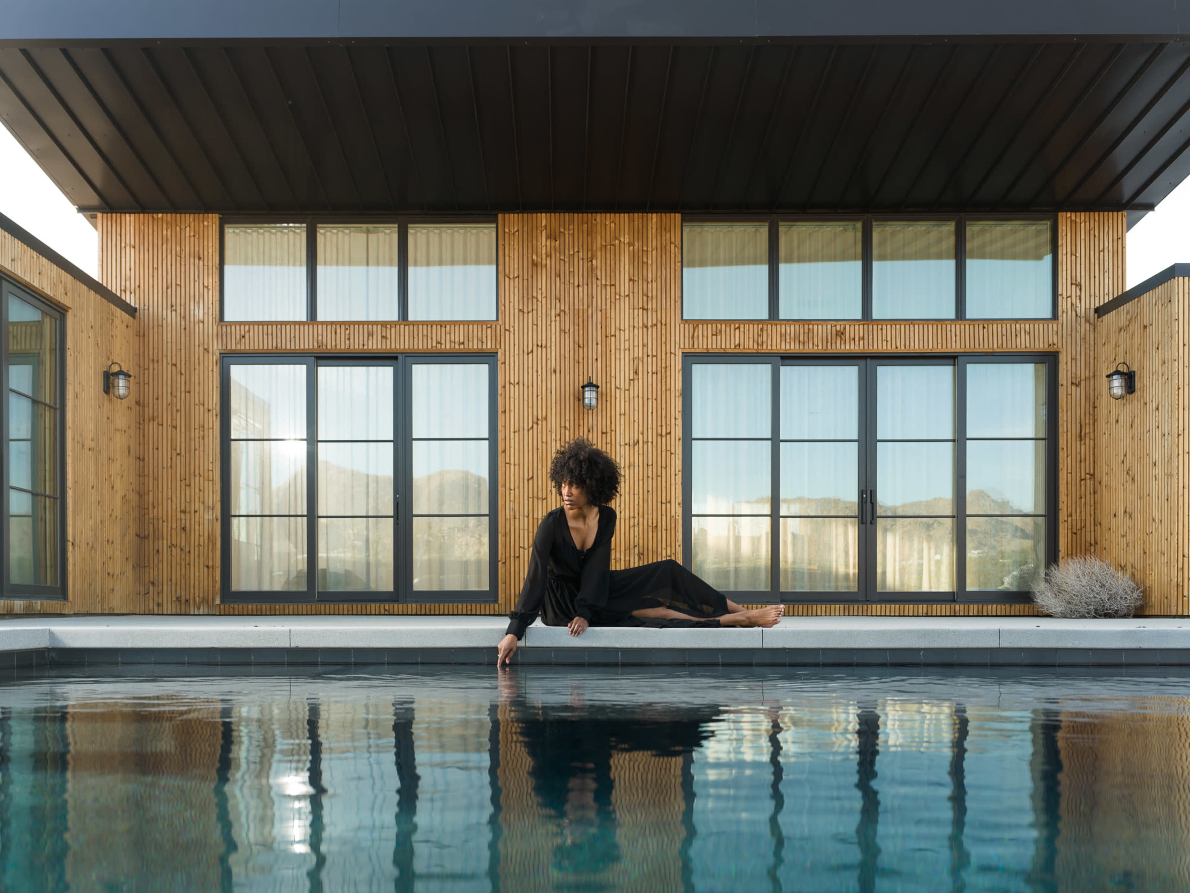 A woman sits on the edge of a pool, reaching down towards the water, in front of a modern wooden building with large windows.