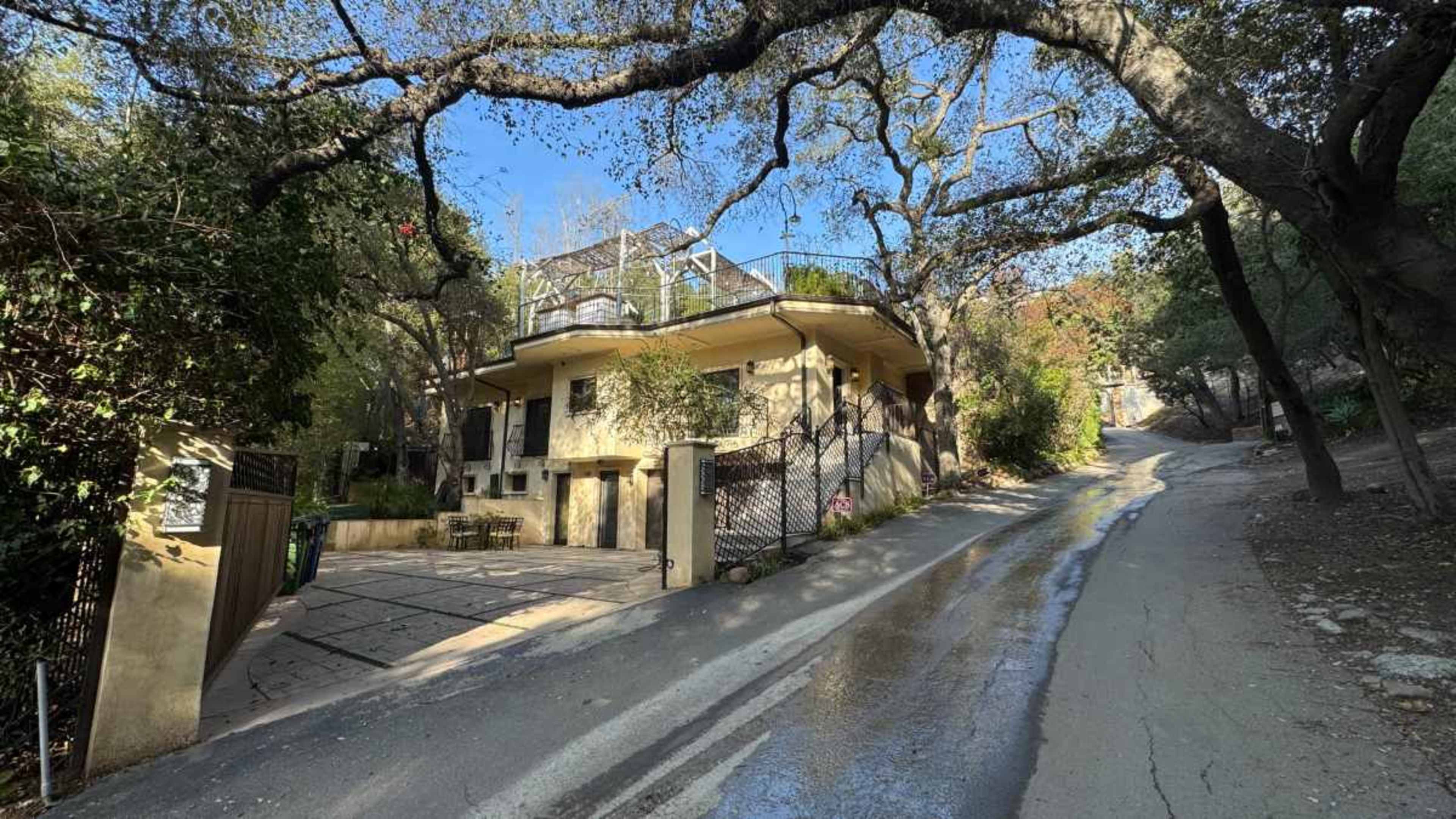 The image shows a house set back from a sloped road, surrounded by trees and foliage.