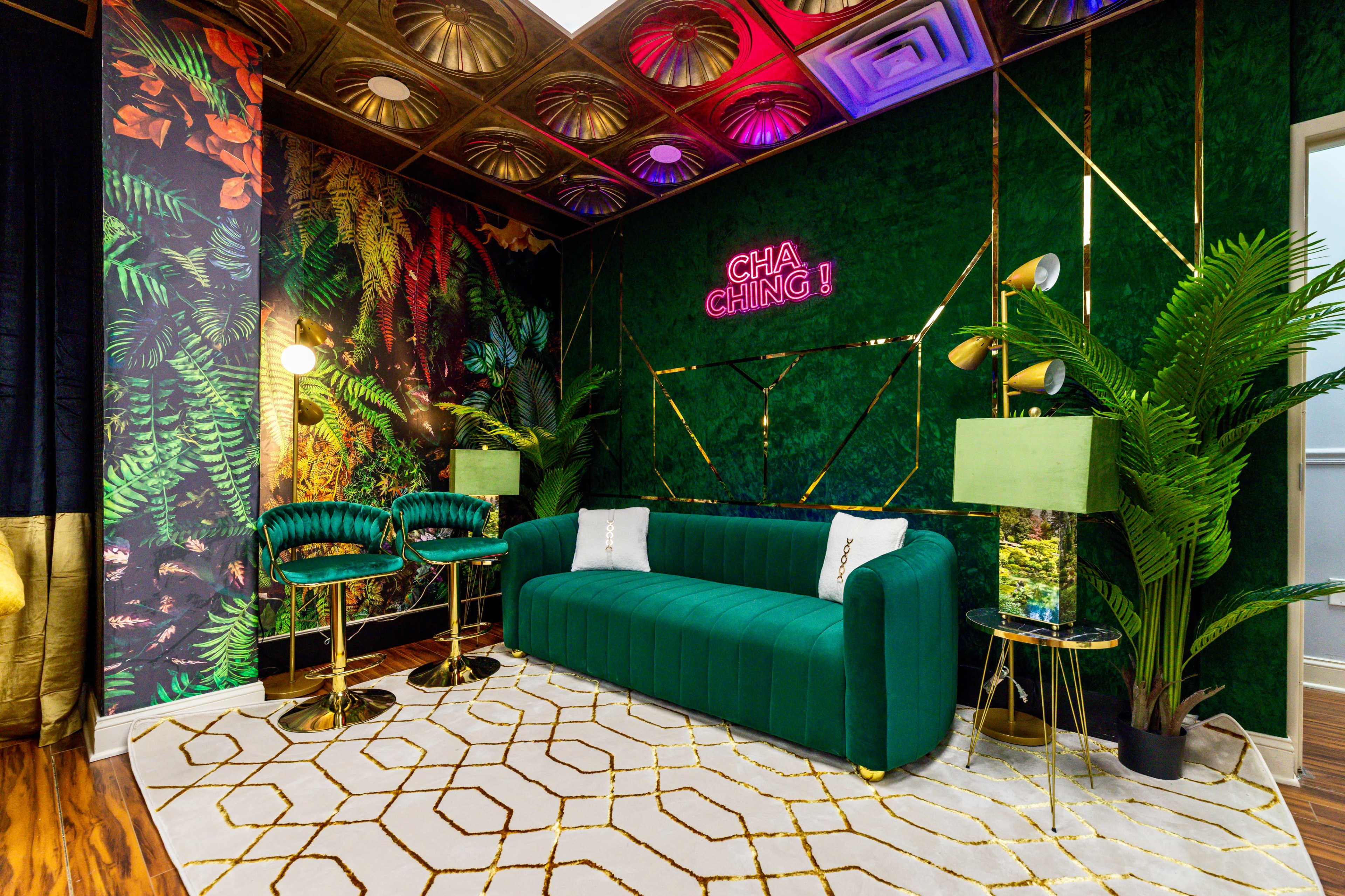 The image shows a stylish lounge area with a green velvet sofa, gold accents, decorative plants, and a vibrant patterned wall featuring tropical foliage.