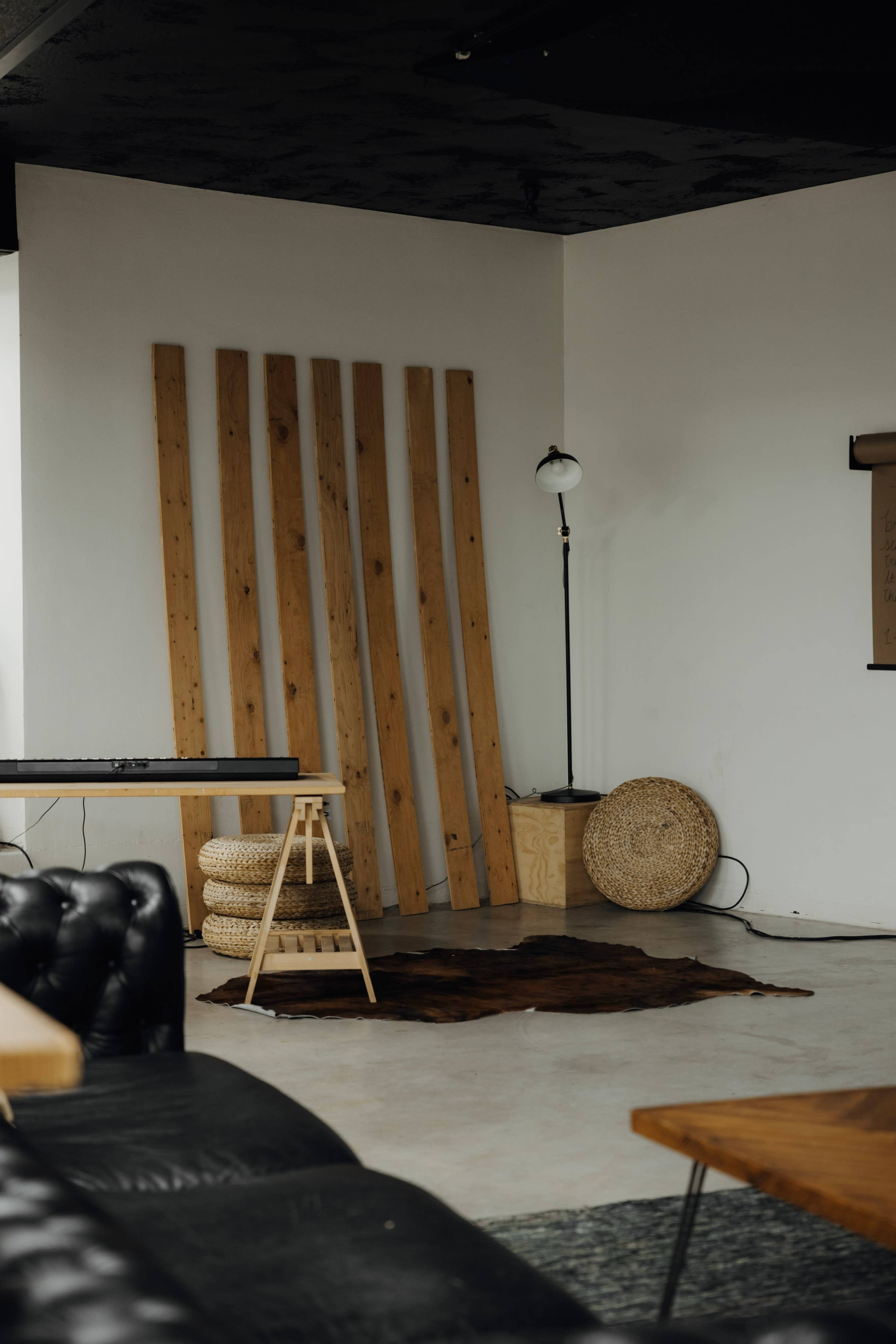 The image shows a minimalist interior space featuring wooden panels leaning against a wall, a standing lamp, a circular woven basket, and a rug on the floor.