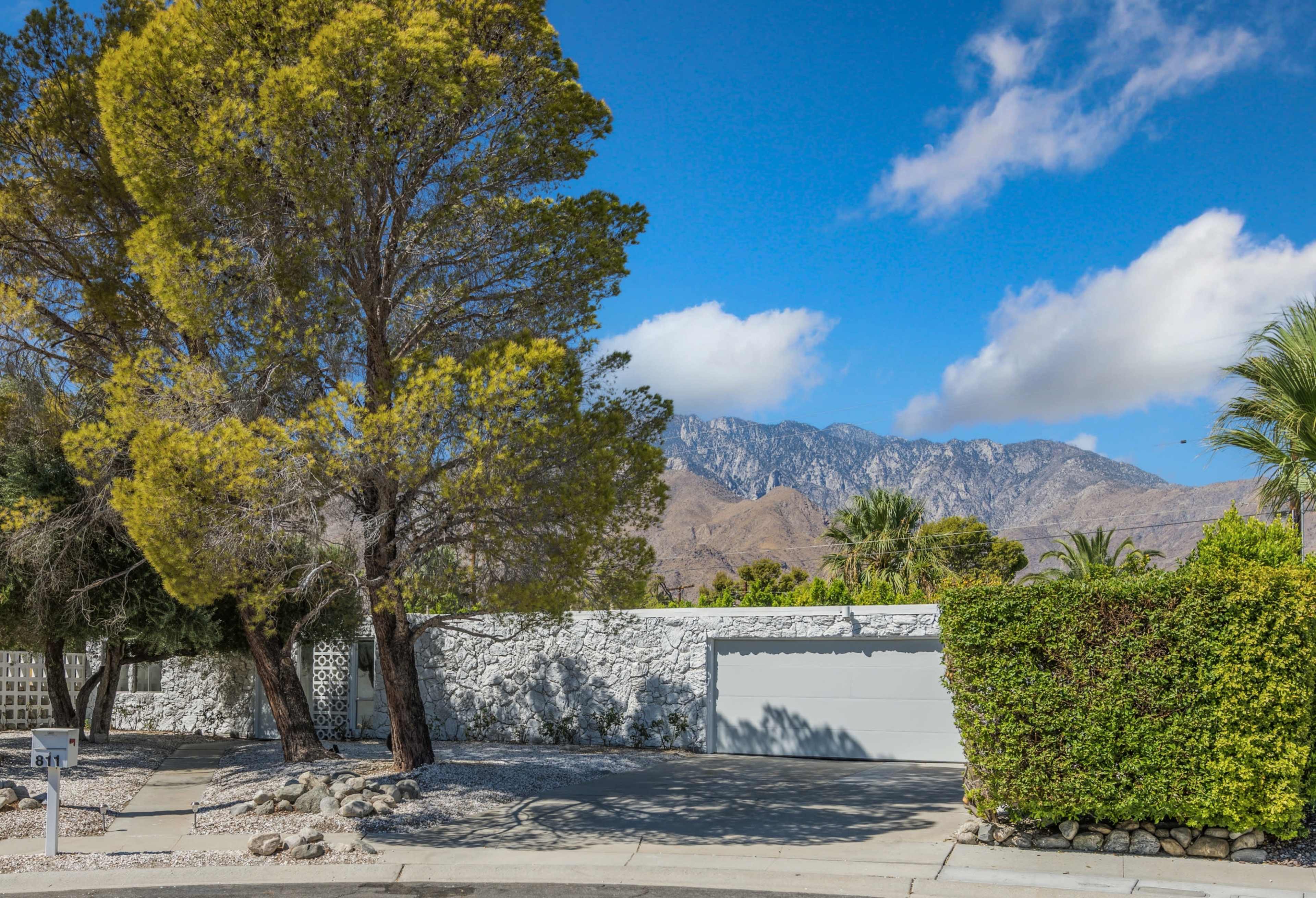A residential property features a white garage door near a landscaped yard with a large tree and a backdrop of mountain hills under a partly cloudy sky.