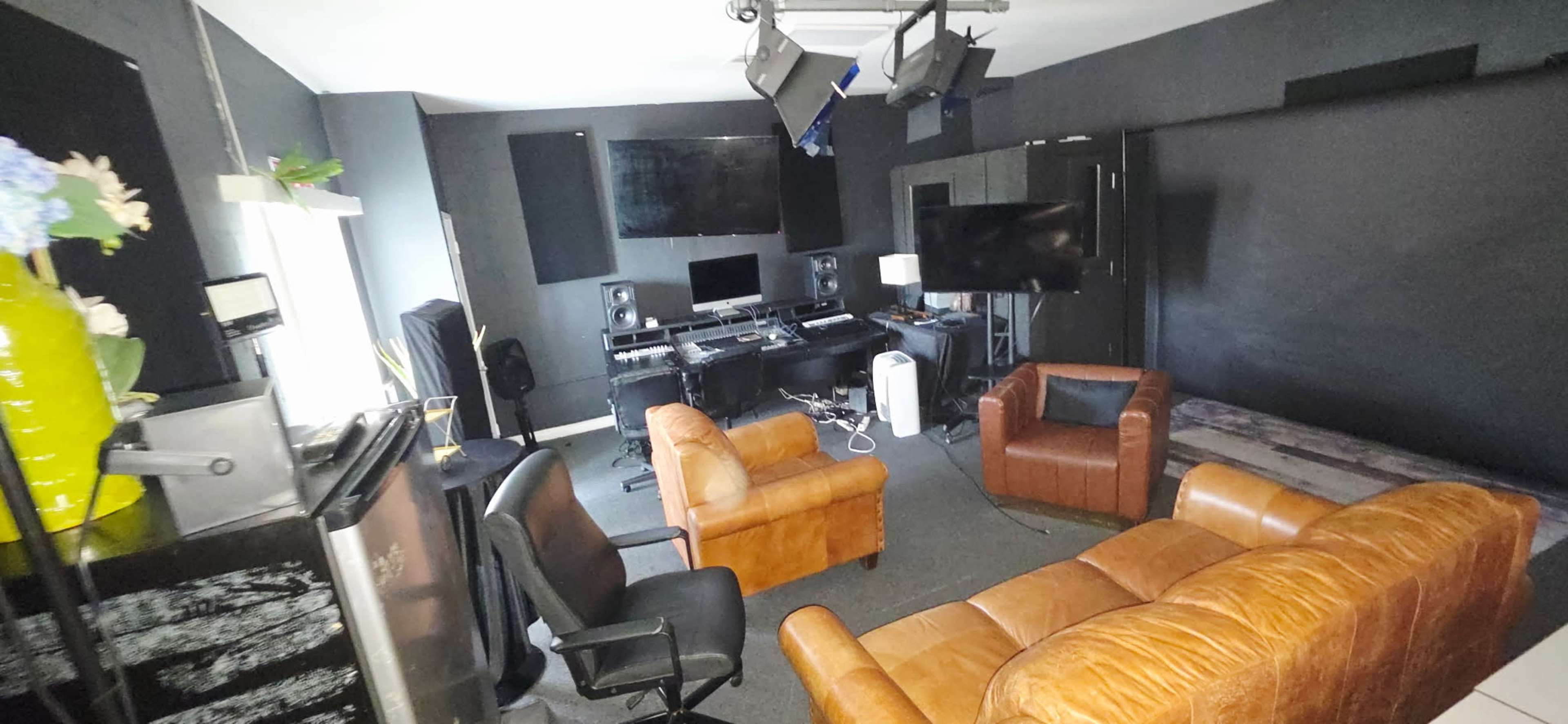 The image shows a modern studio or lounge area featuring two leather sofas, a black office chair, and multiple screens and audio equipment set against dark walls.