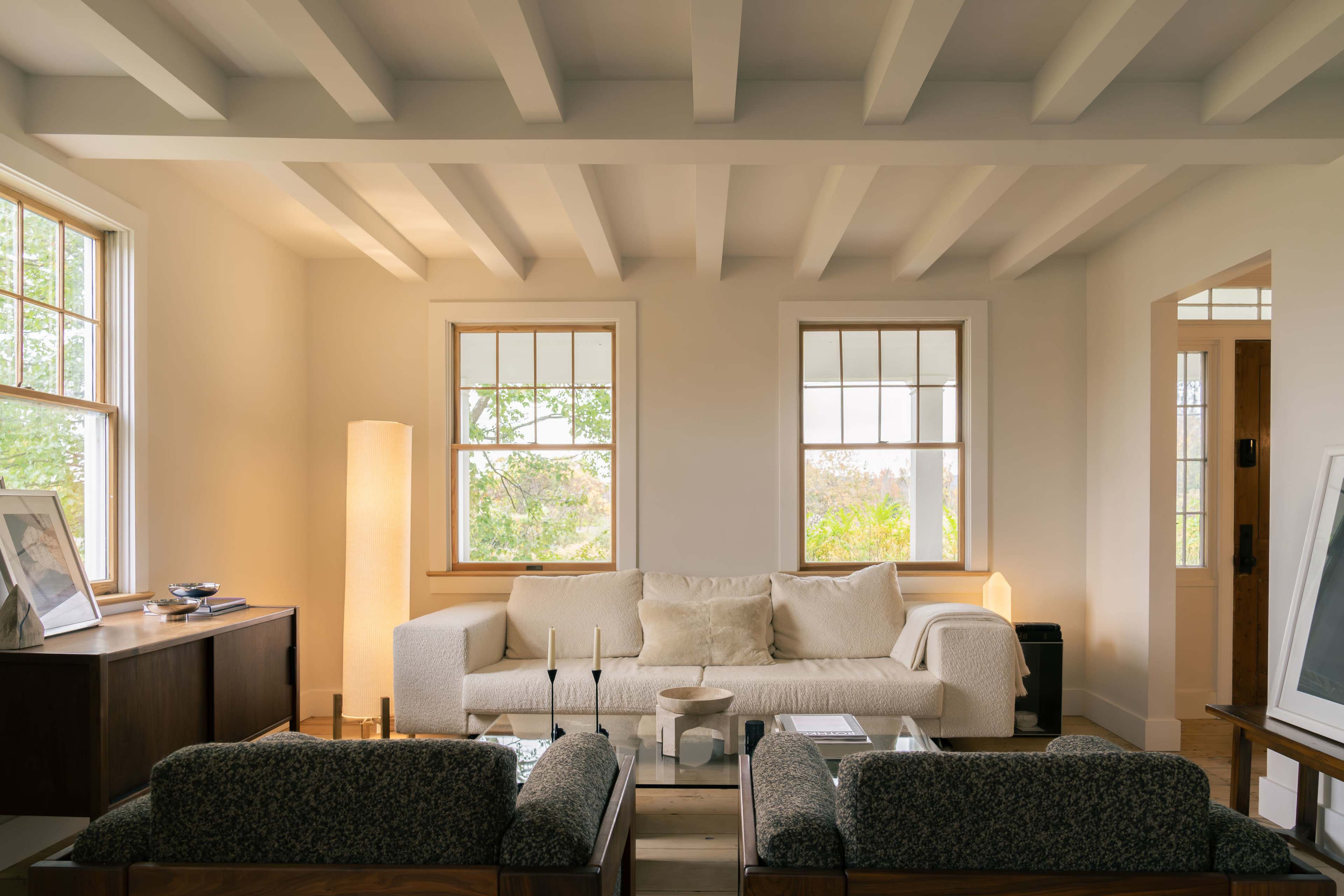 The living room features a white sofa, wooden furniture, and large windows that allow natural light to fill the space.