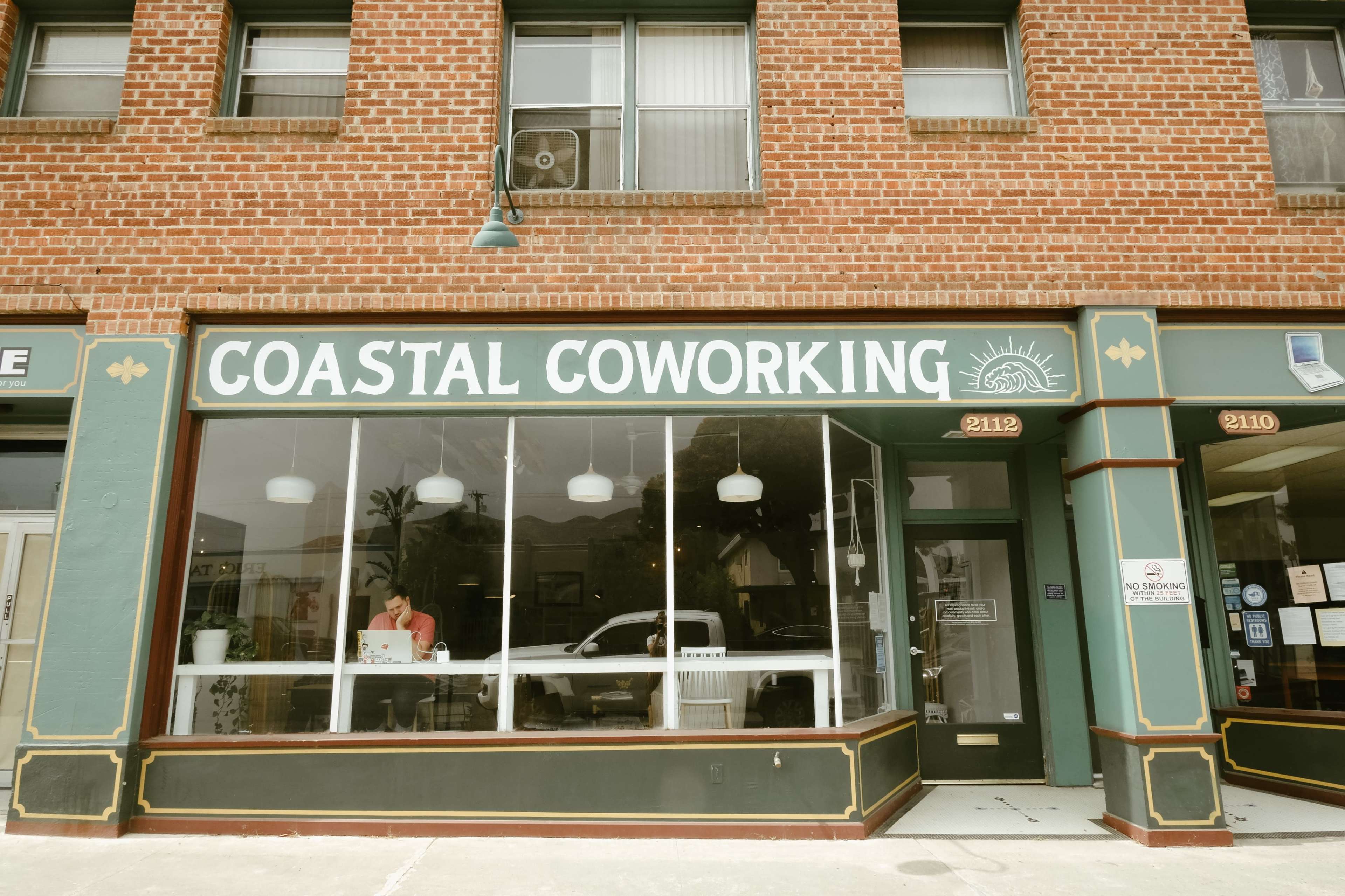 The exterior of a brick building with large windows displaying a workspace labeled "Coastal Coworking."