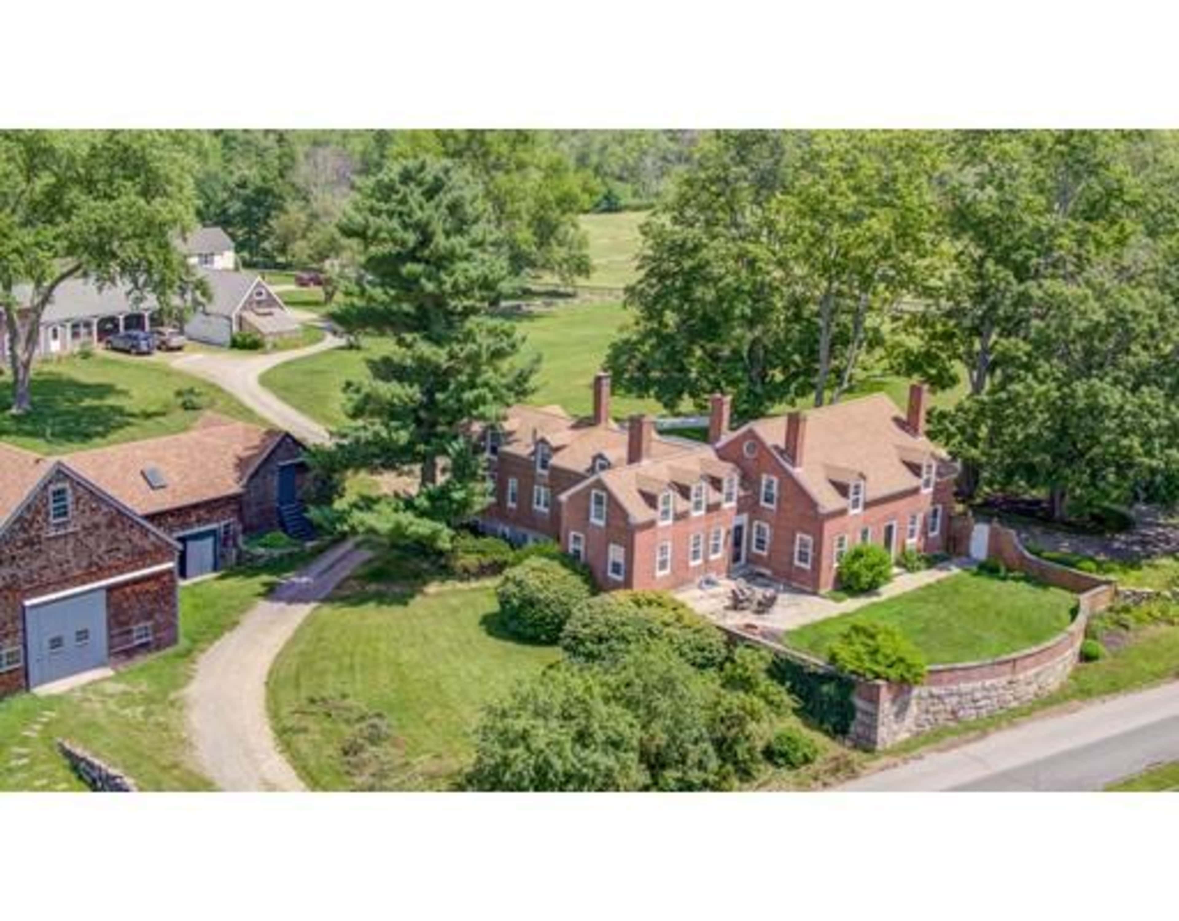 A large brick house with multiple chimneys is situated in a lush, green landscape, surrounded by trees and outbuildings.
