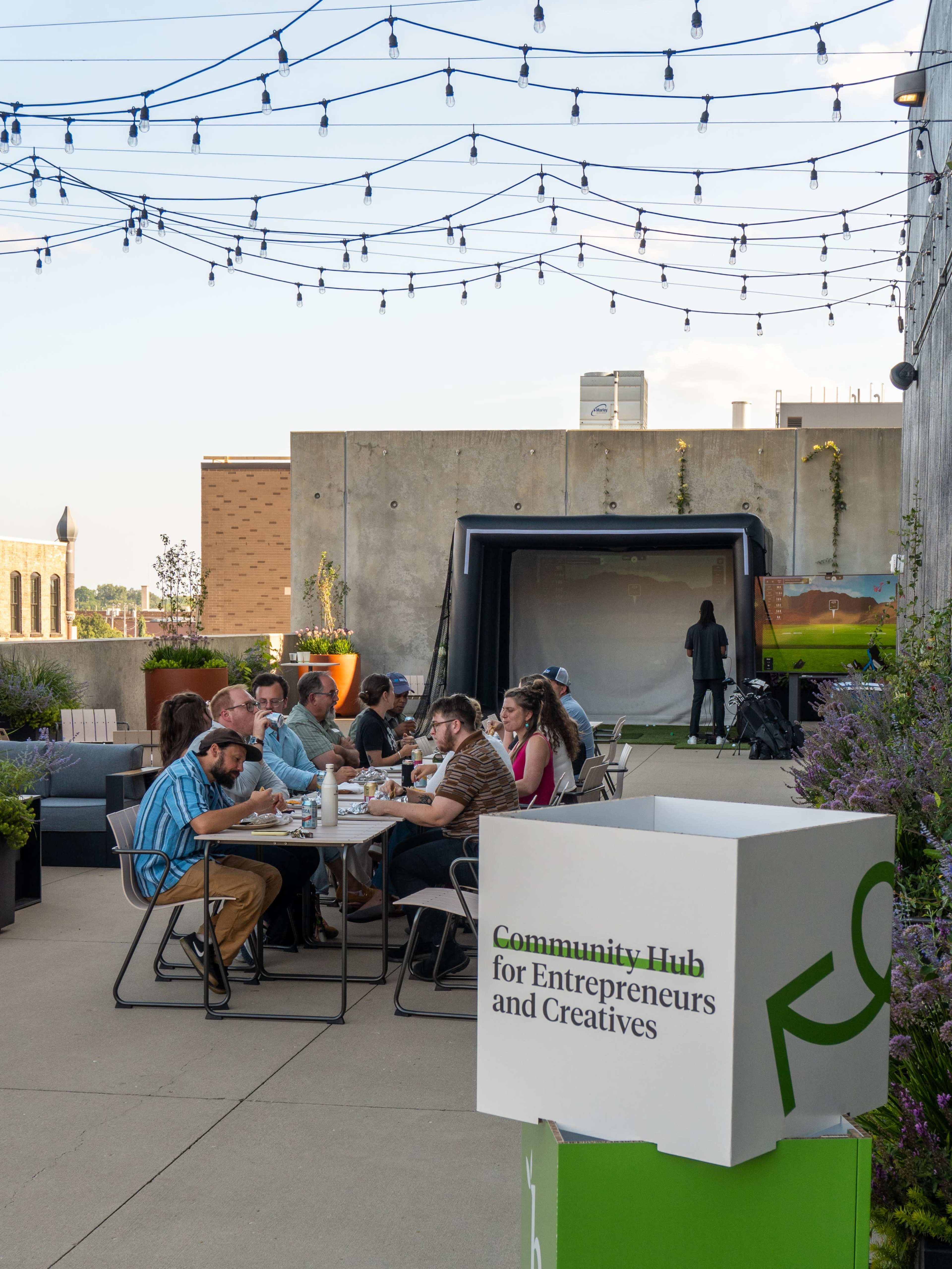 A group of people is seated at a long table on a rooftop terrace, dining together under string lights, with a backdrop featuring greenery and a projection screen.
