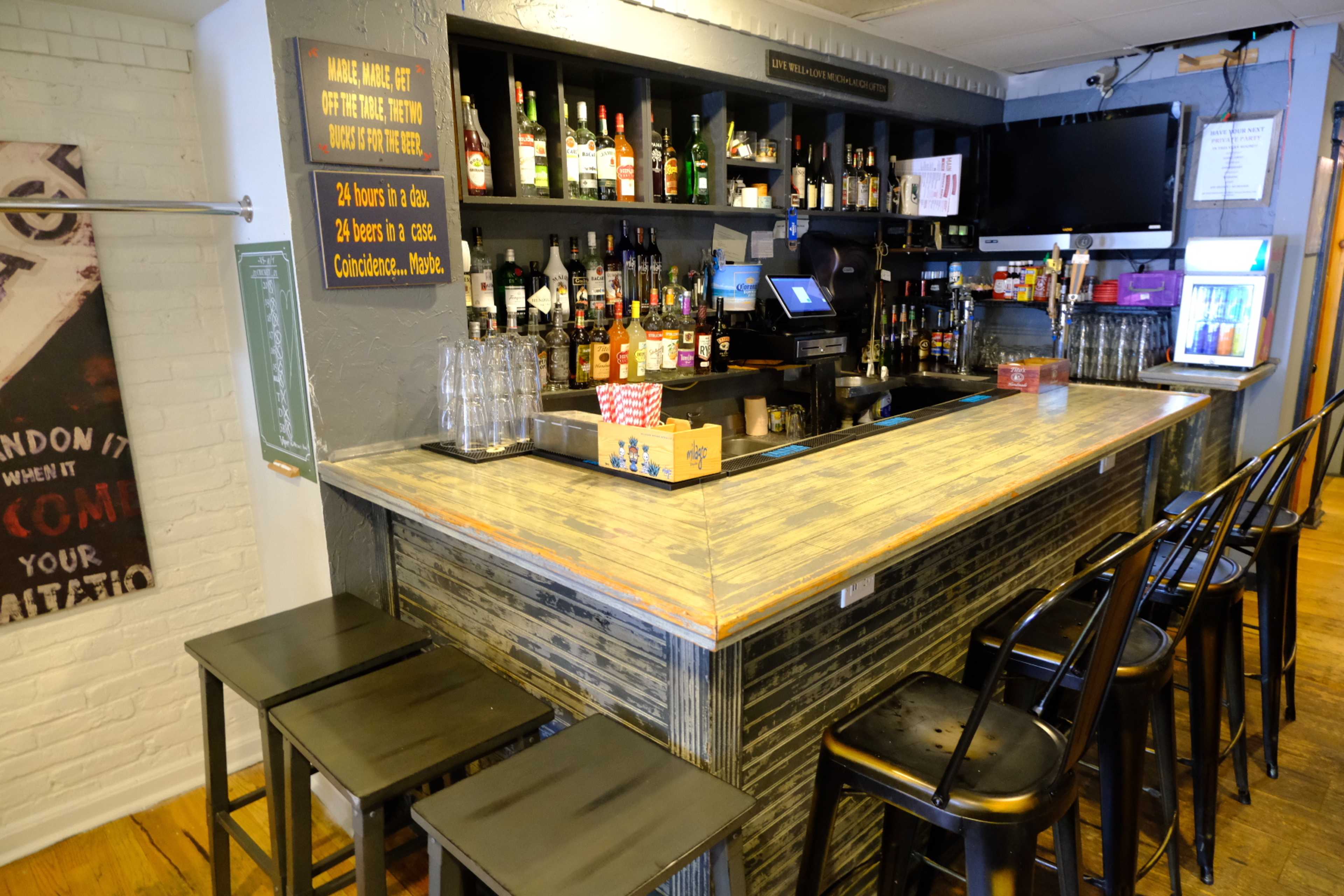 A bar area with a polished countertop, shelves stocked with various liquor bottles, and several bar stools arranged in front.
