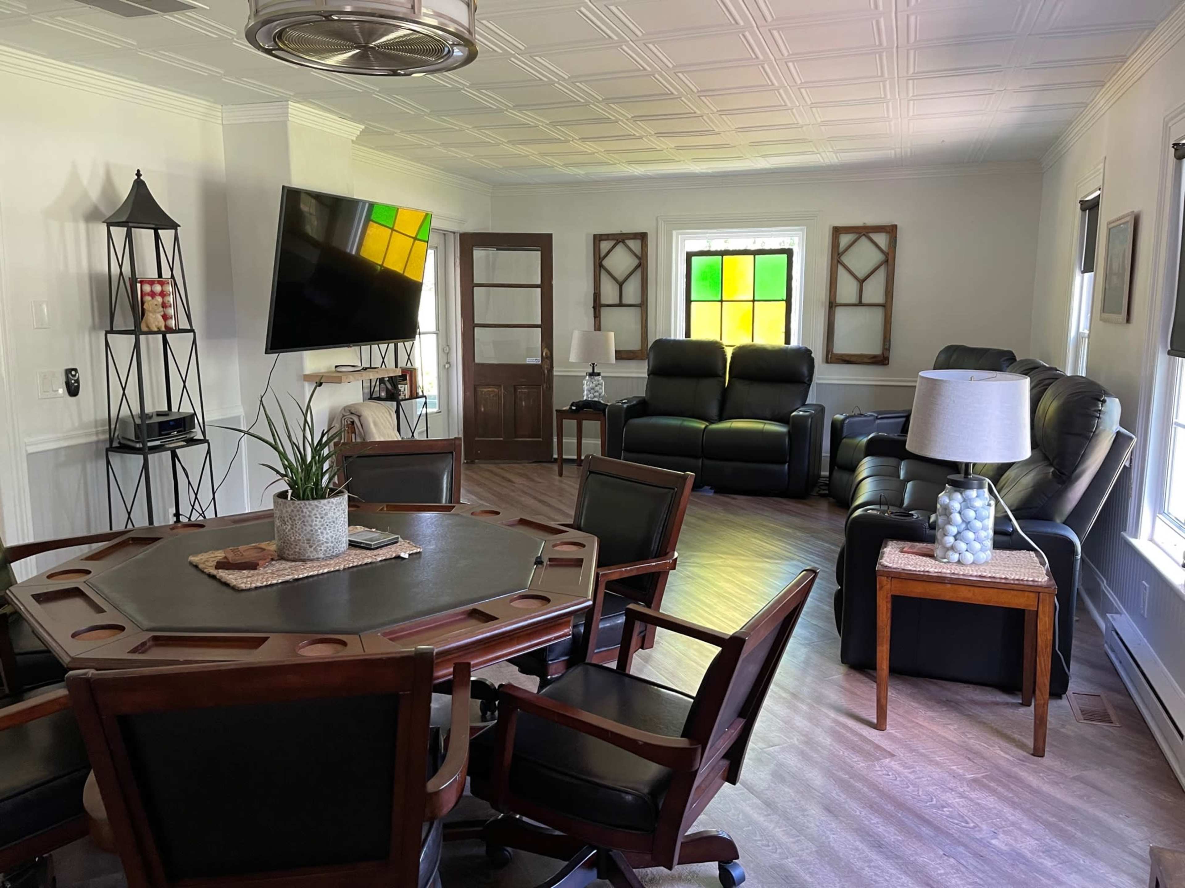 A living room with a dining table and chairs, a large television mounted on the wall, and two recliners situated to the right, along with a coffee table.
