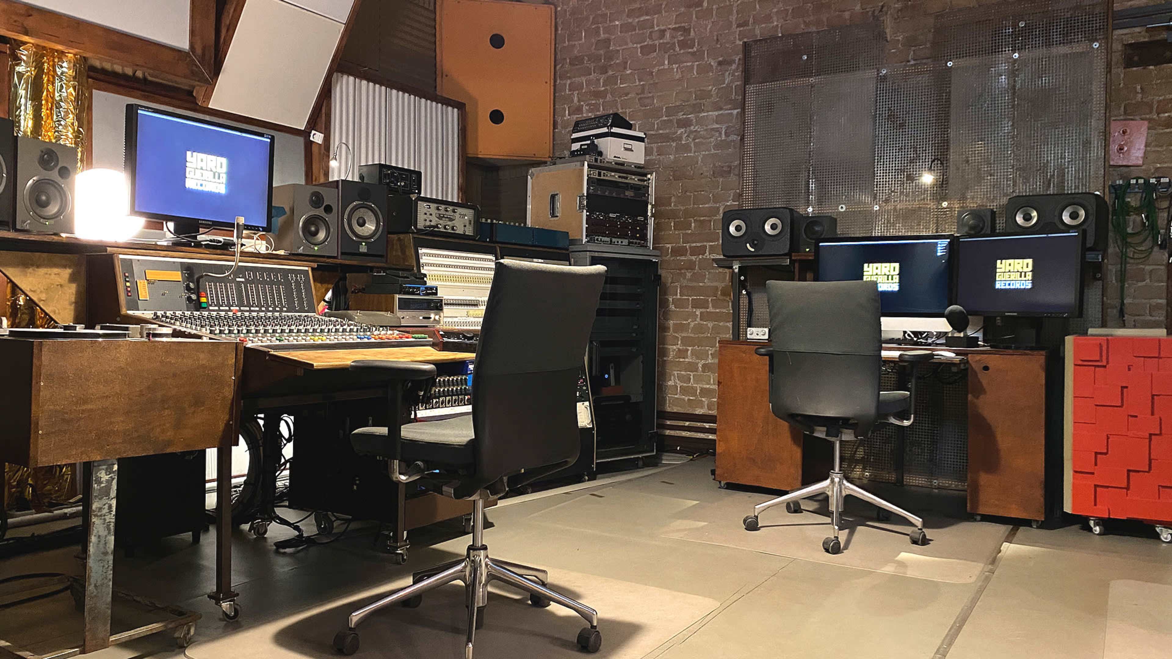 The image shows a music studio with a mixing console, two computer workstations, and various audio equipment arranged in a room with brick walls.