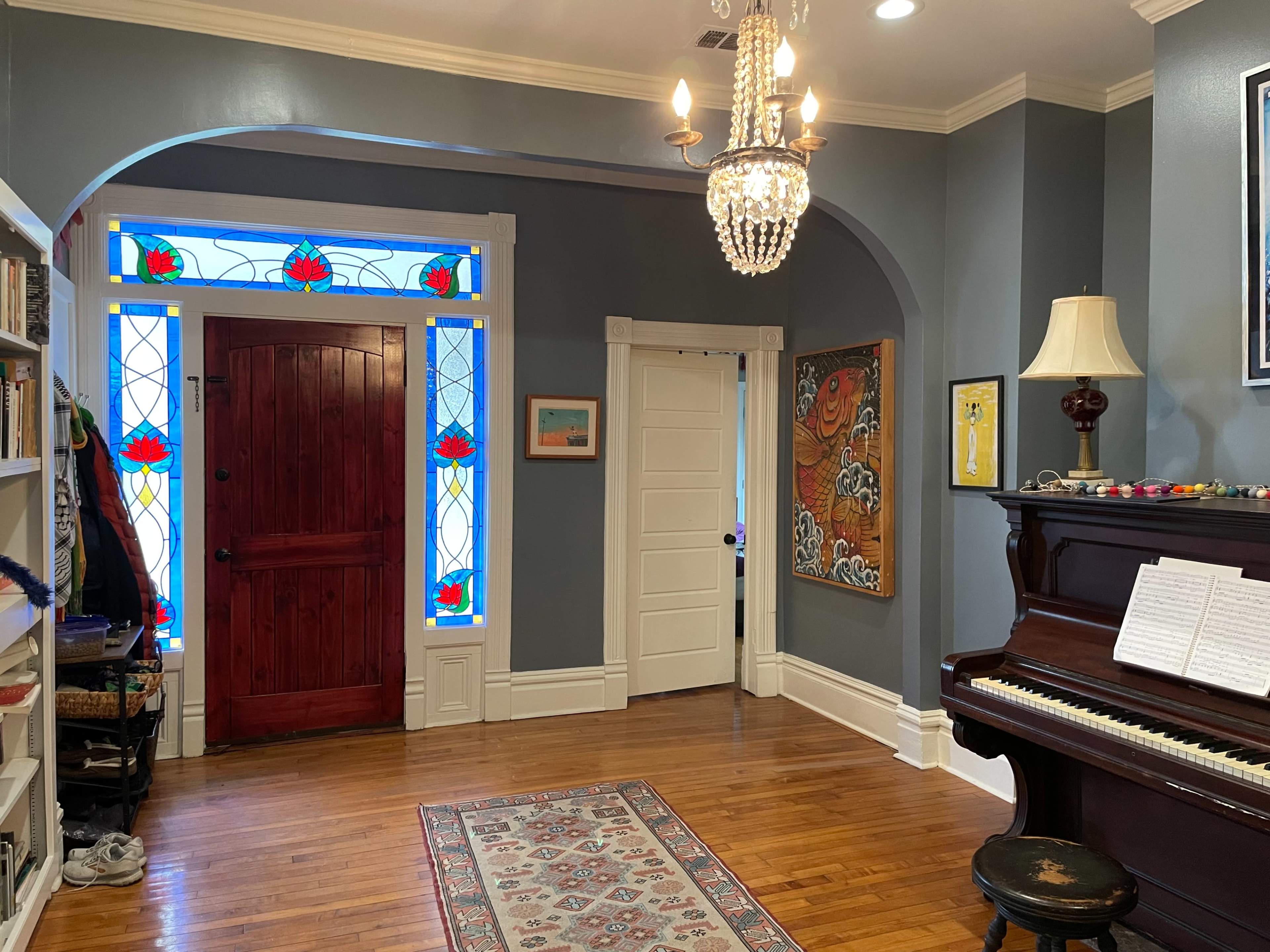 A foyer features stained glass windows, a chandelier, a piano, and a wooden door.
