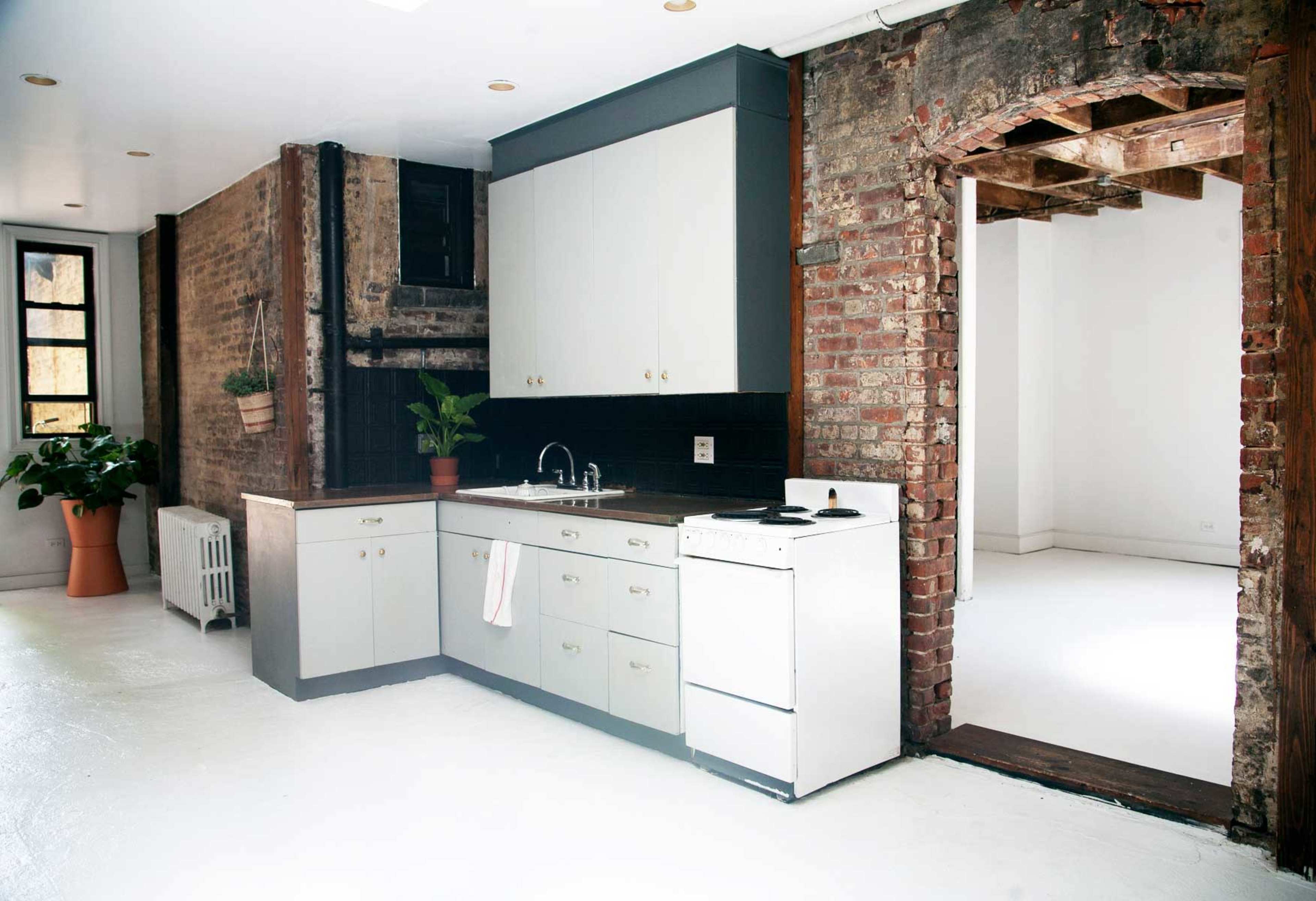 A kitchen with minimalist cabinetry, a white stove, and exposed brick walls, leading to an empty, light-filled room.
