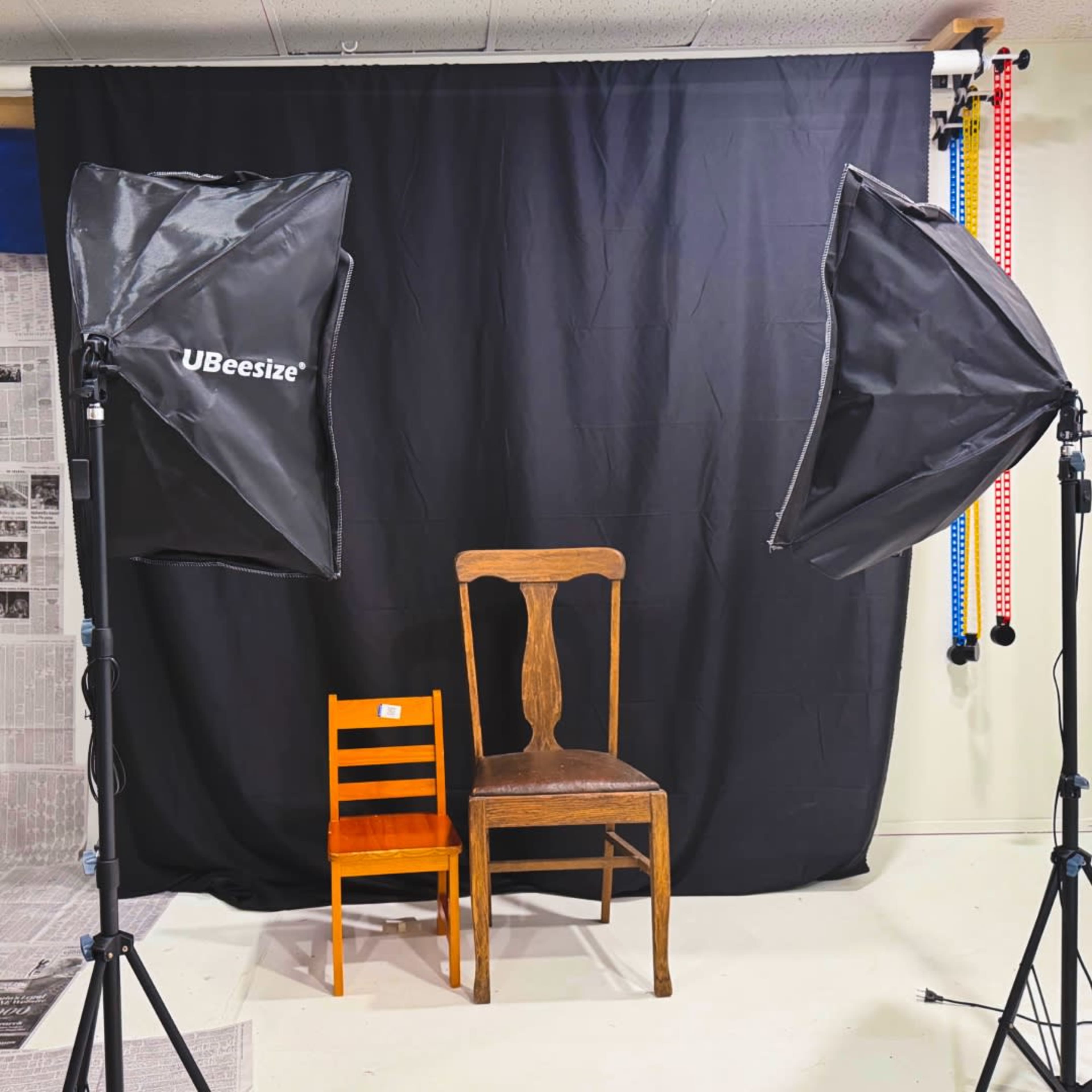 The image shows two chairs, one orange and small, positioned next to a larger wooden chair, in front of a black backdrop with softbox lighting on either side.