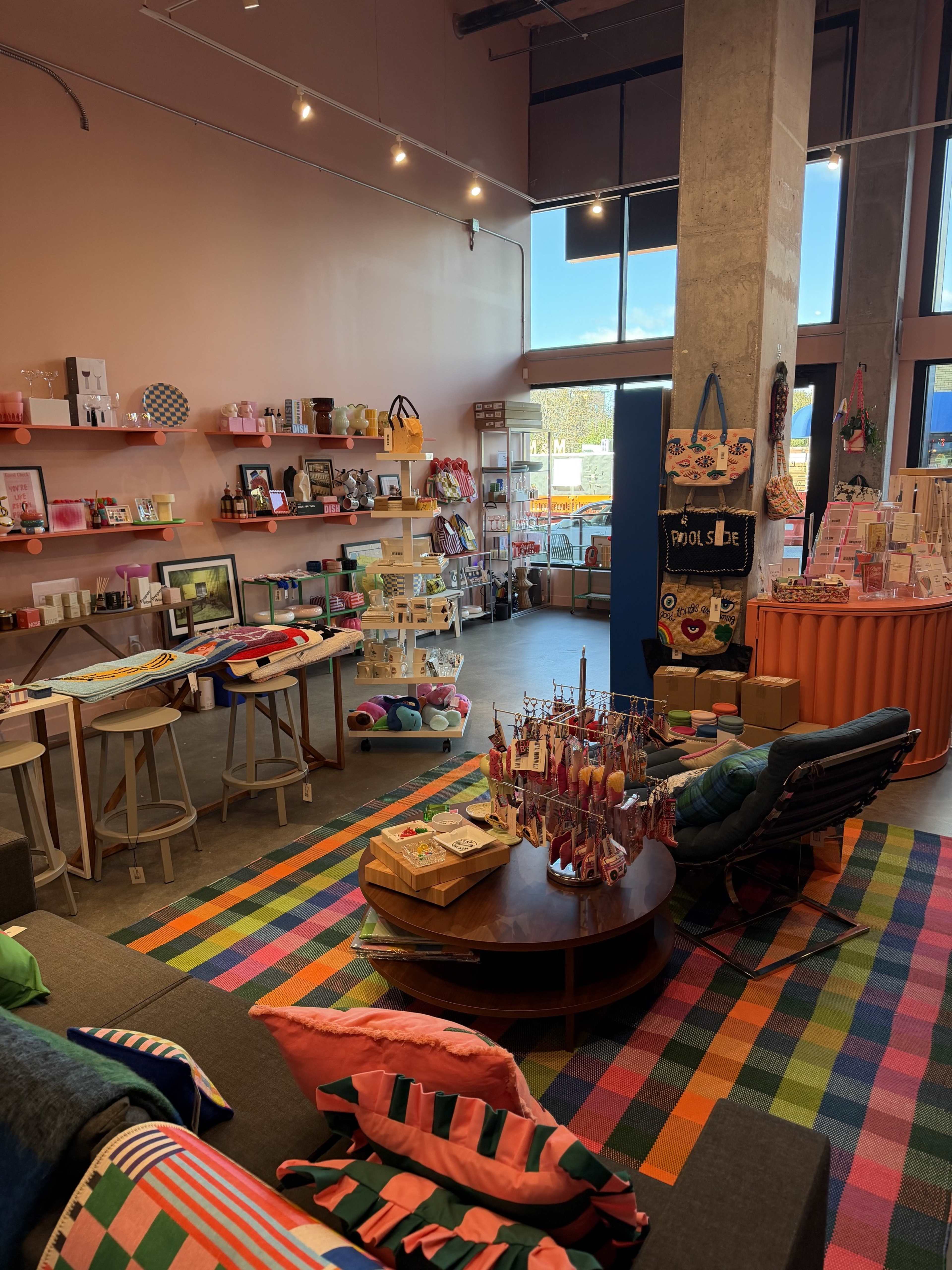 The image shows a colorful retail space featuring shelves filled with various products, a seating area with striped cushions, and large windows allowing natural light to enter.