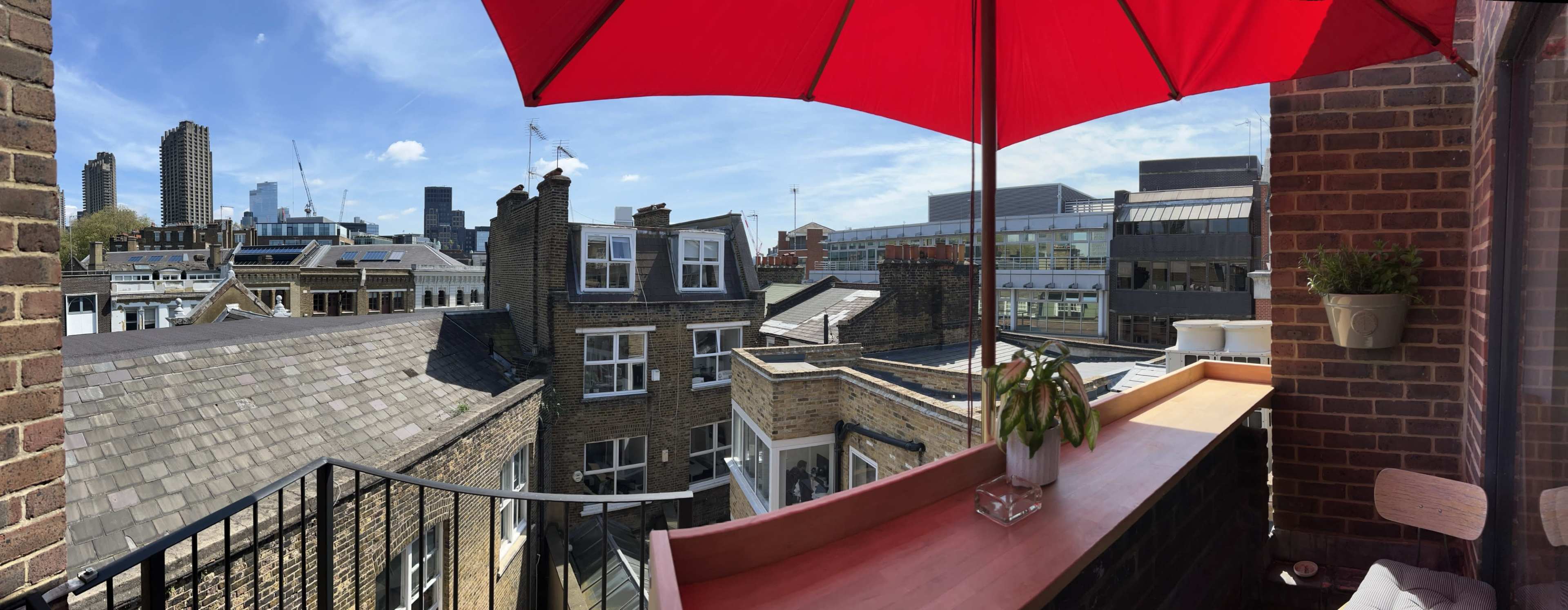 A red umbrella shades a balcony overlooking the rooftops and skyline of a city on a clear day.