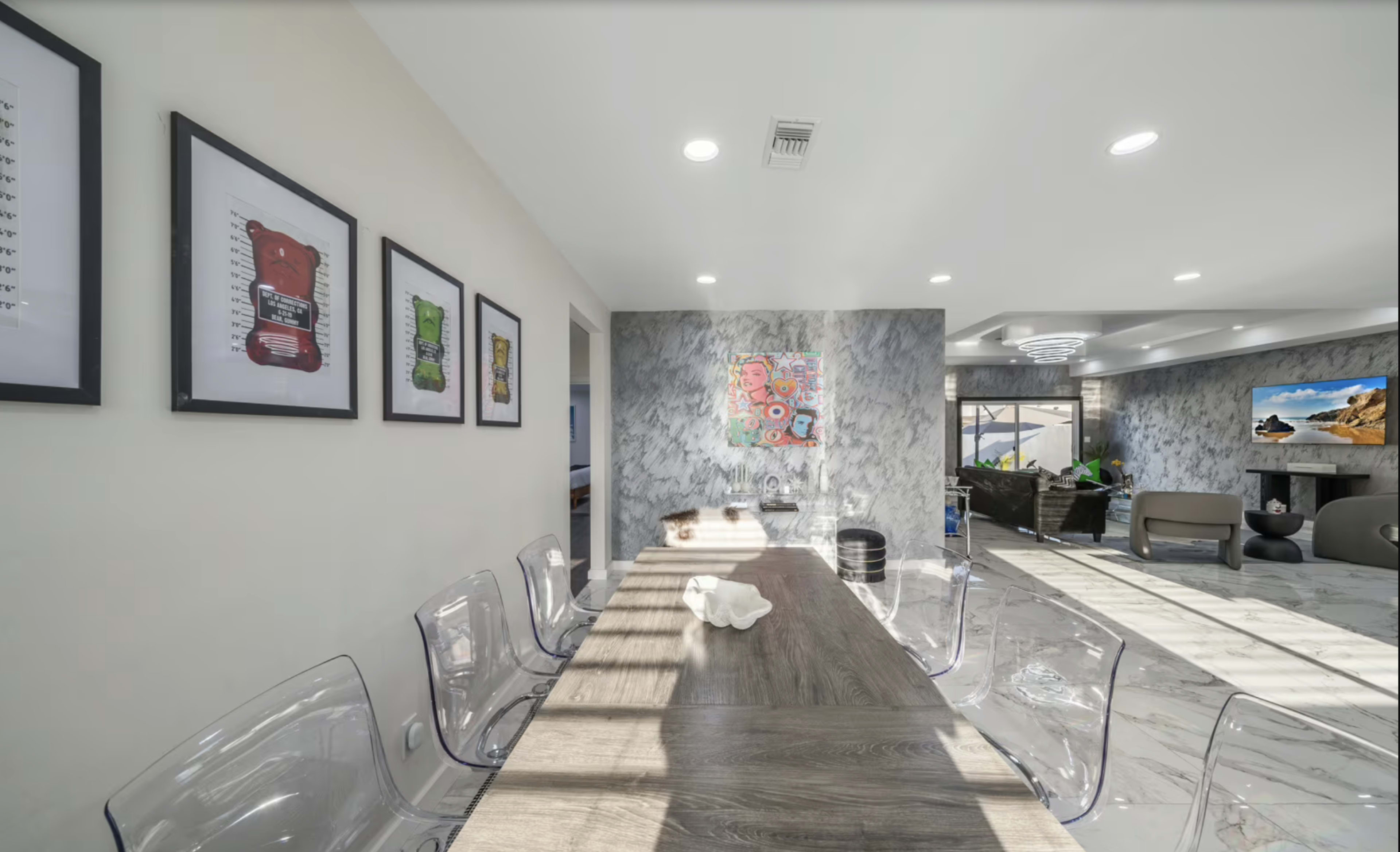 A modern dining area features a long wooden table surrounded by transparent chairs, with artwork displayed on the walls and a spacious, well-lit living area visible in the background.