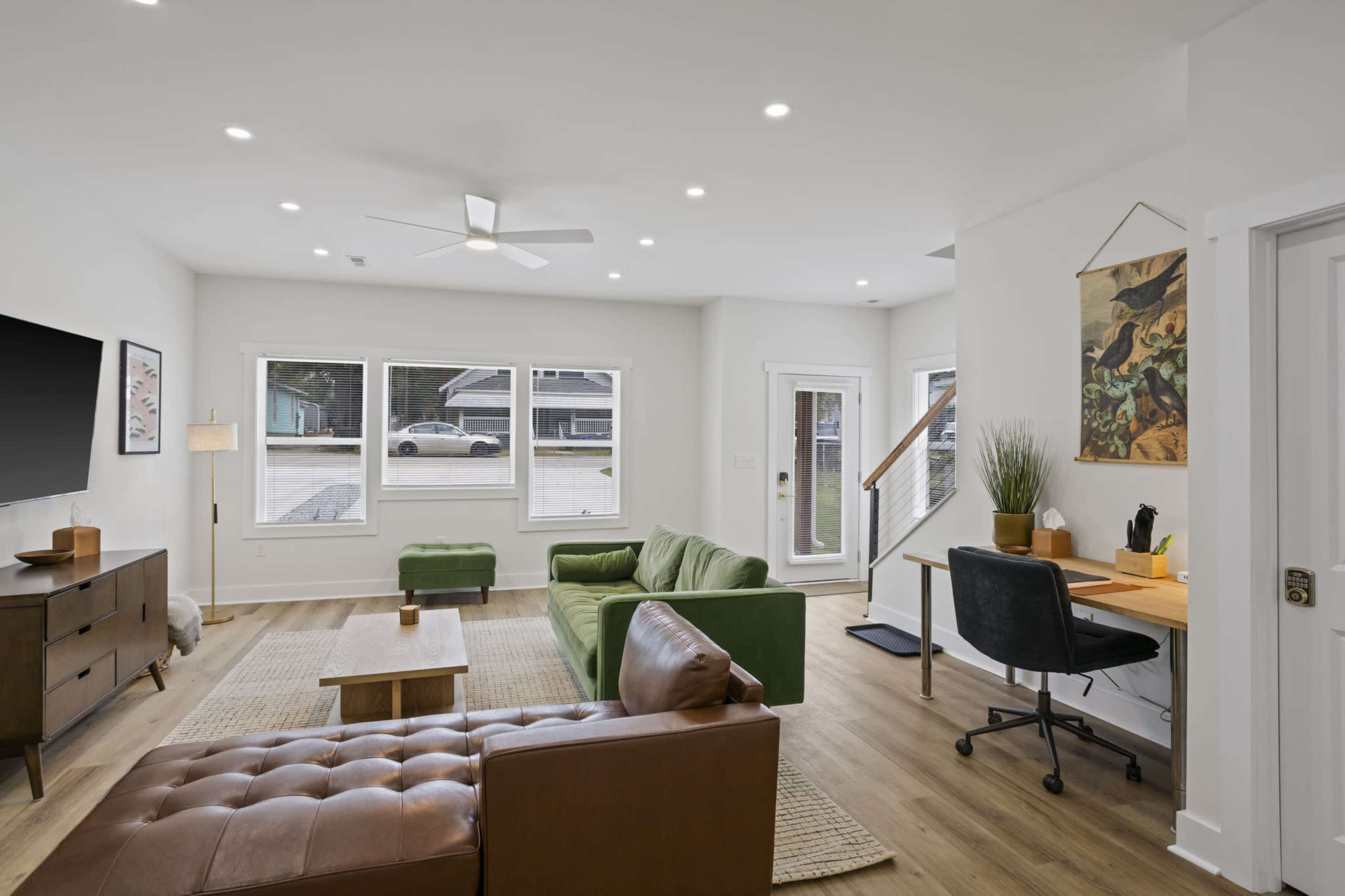 The image shows a modern living room with green and brown furniture, large windows, and a small workspace in one corner.