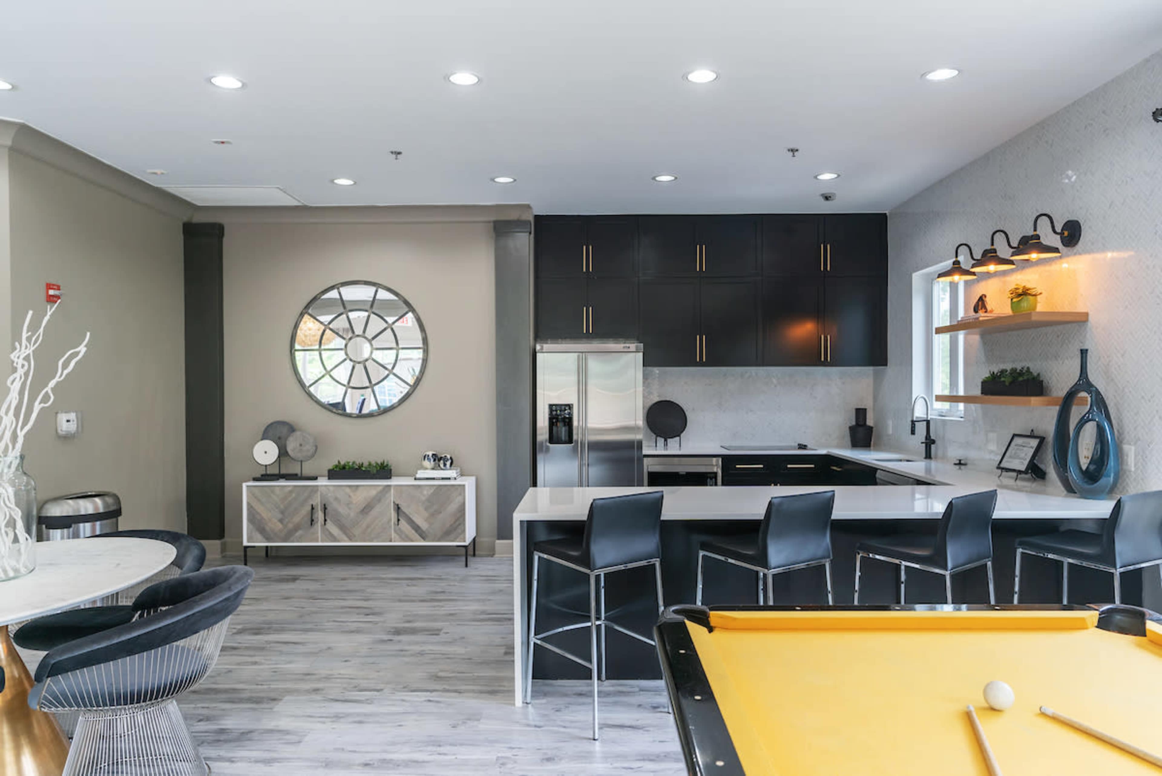 A modern kitchen and dining area featuring black cabinetry, a large round mirror, and a pool table in the foreground.