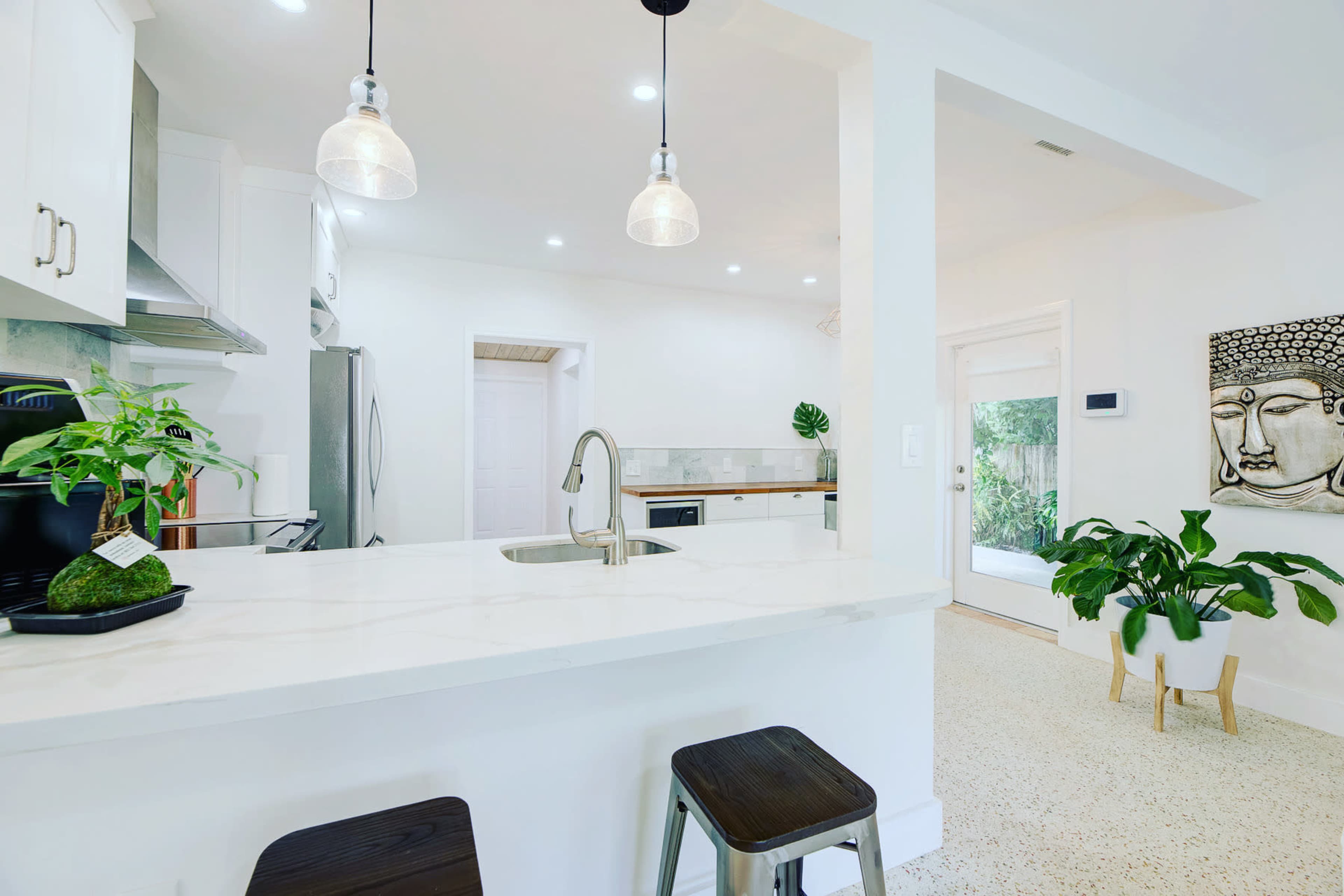 A modern kitchen features a white marble countertop, stainless steel appliances, and a small dining area with plants and an artwork on the wall.