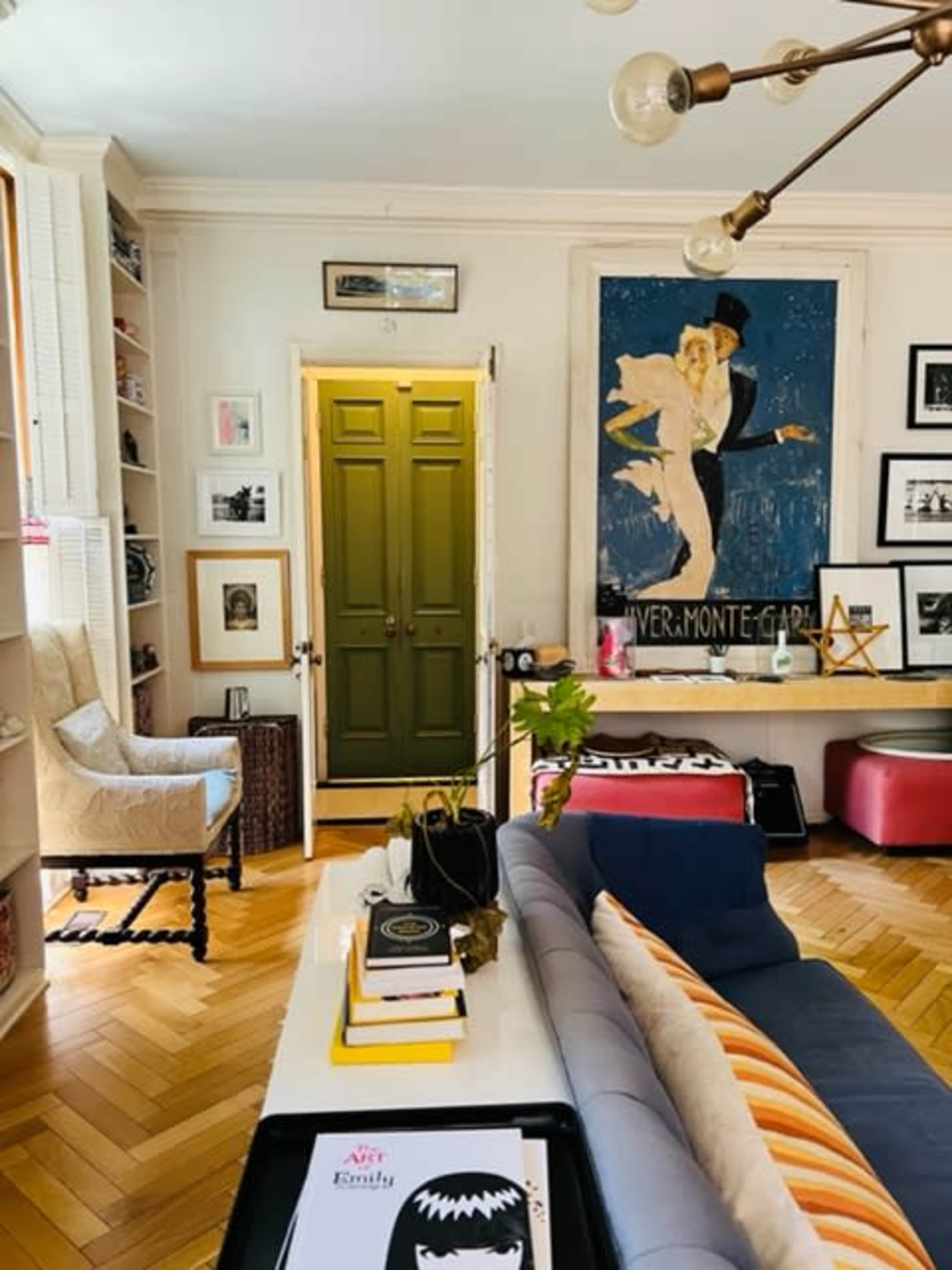 The scene shows a cozy living room with a green door, a seating area with a blue and white couch, shelves filled with books and decor, and a vintage-style poster on the wall.