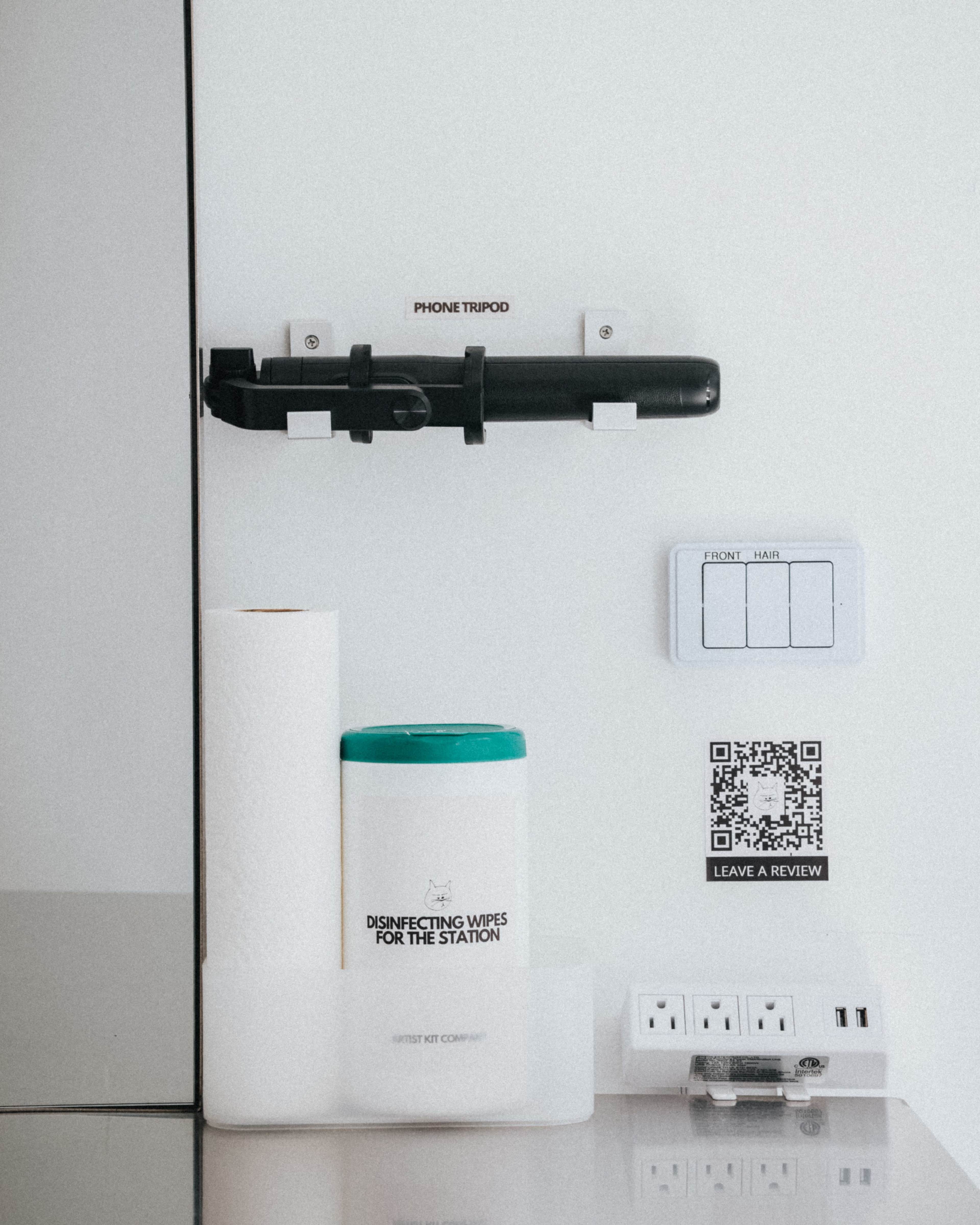 The image shows a wall with a phone tripod holder, a container of disinfecting wipes, a paper towel roll, a QR code, and multiple electrical outlets.