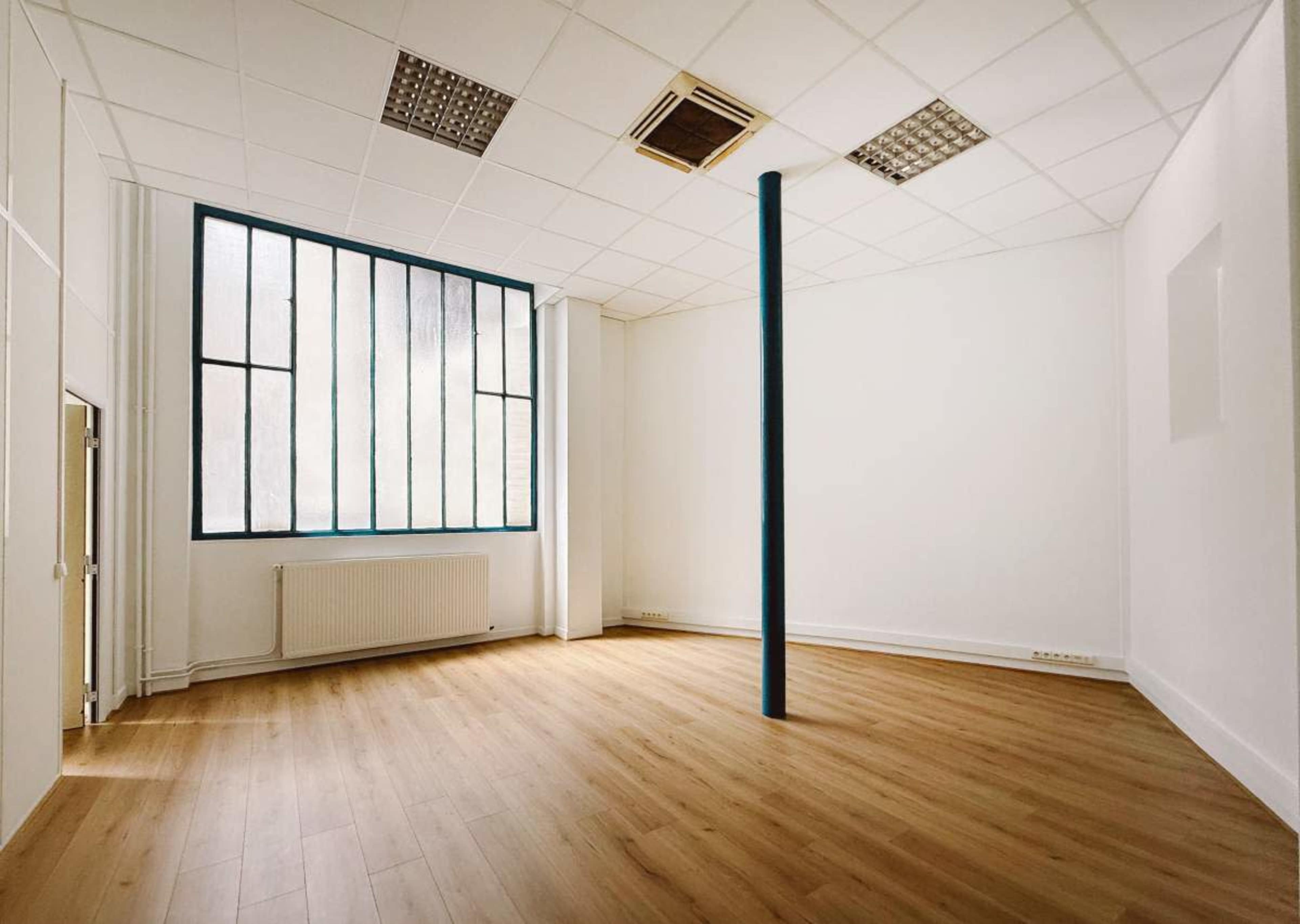 The image shows an empty room with wooden flooring, a vertical blue column, and large windows allowing natural light.
