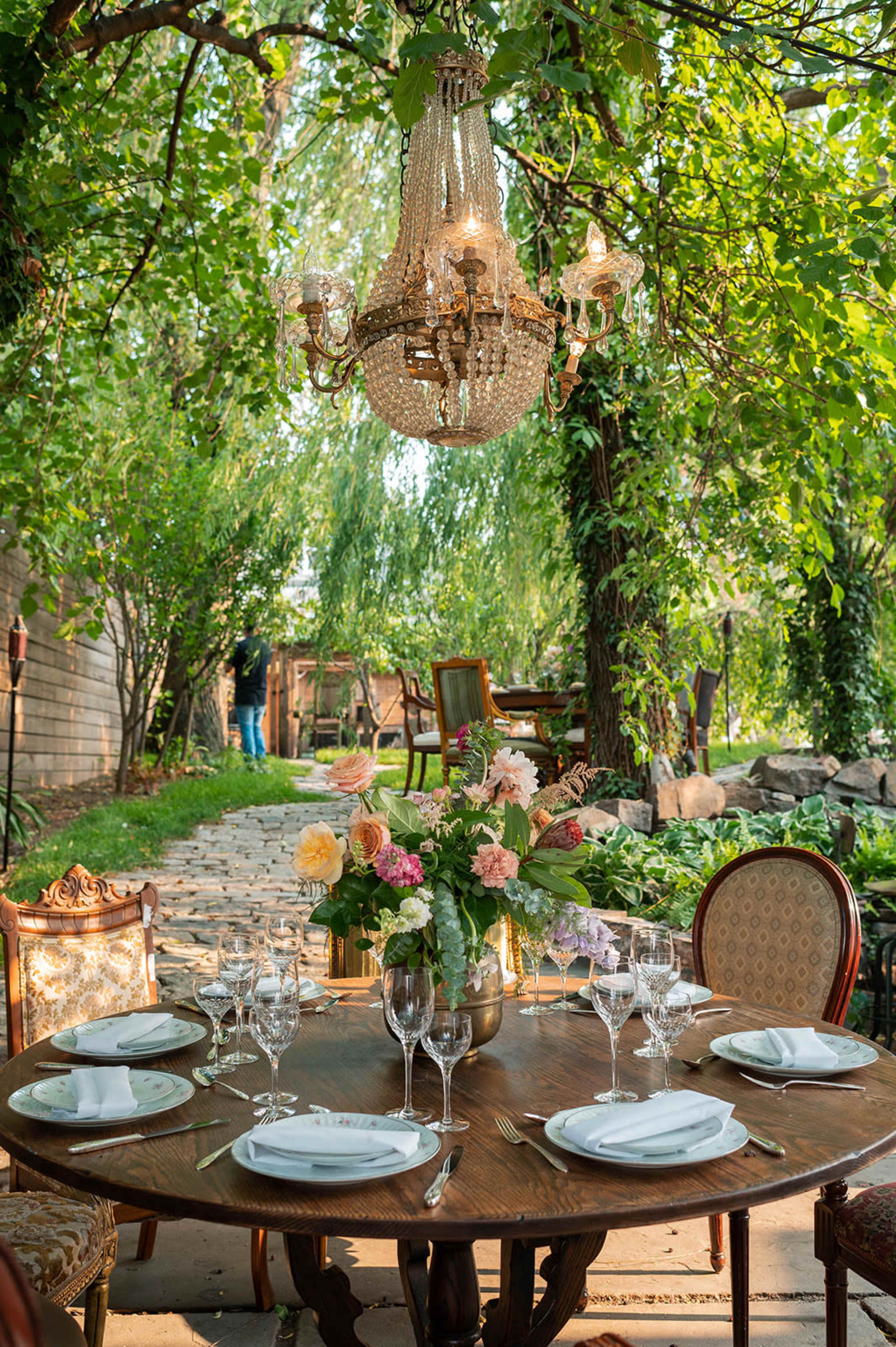 A beautifully set outdoor dining table with a chandelier overhead, surrounded by lush greenery and flowers.