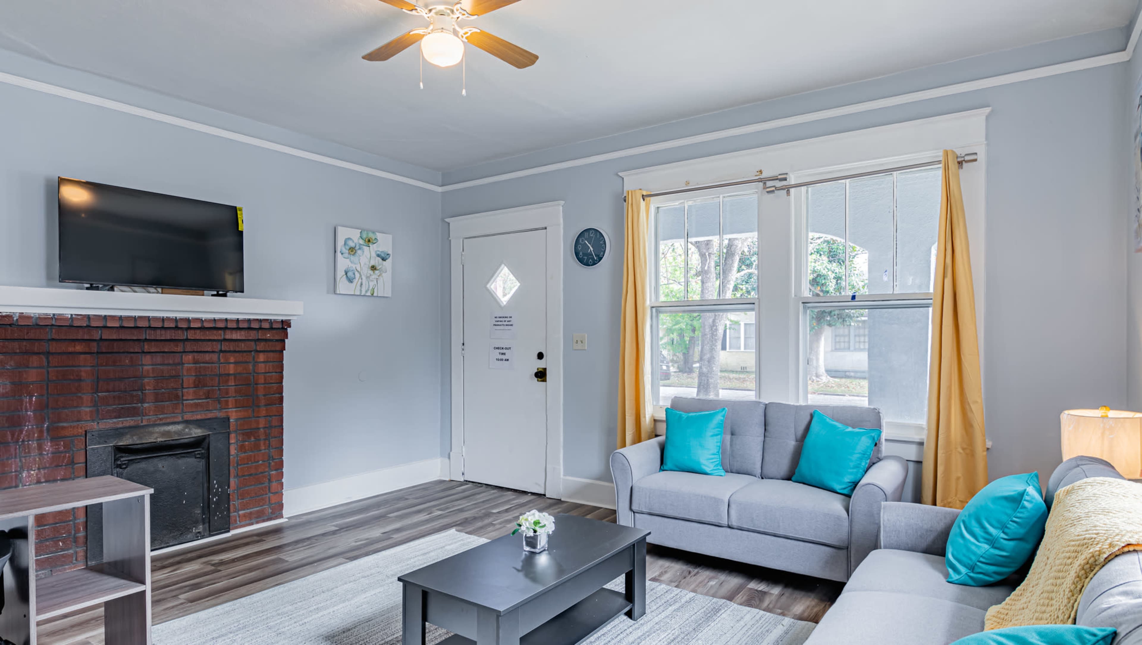 The living room features a gray sofa with teal cushions, a coffee table, a wall-mounted TV, and large windows with yellow curtains.