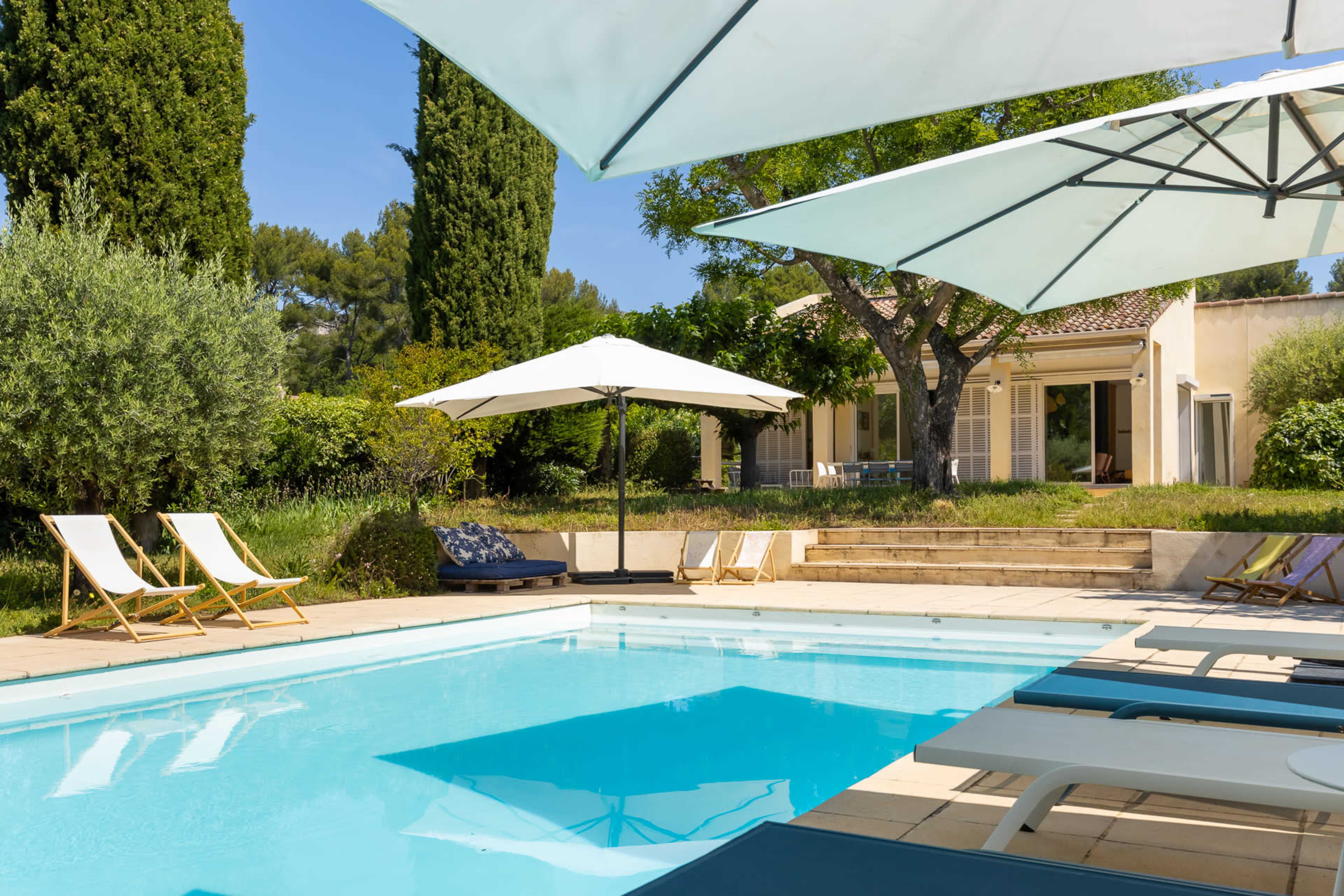 A clear swimming pool is surrounded by lounge chairs and large umbrellas, with a villa and landscaped greenery visible in the background.