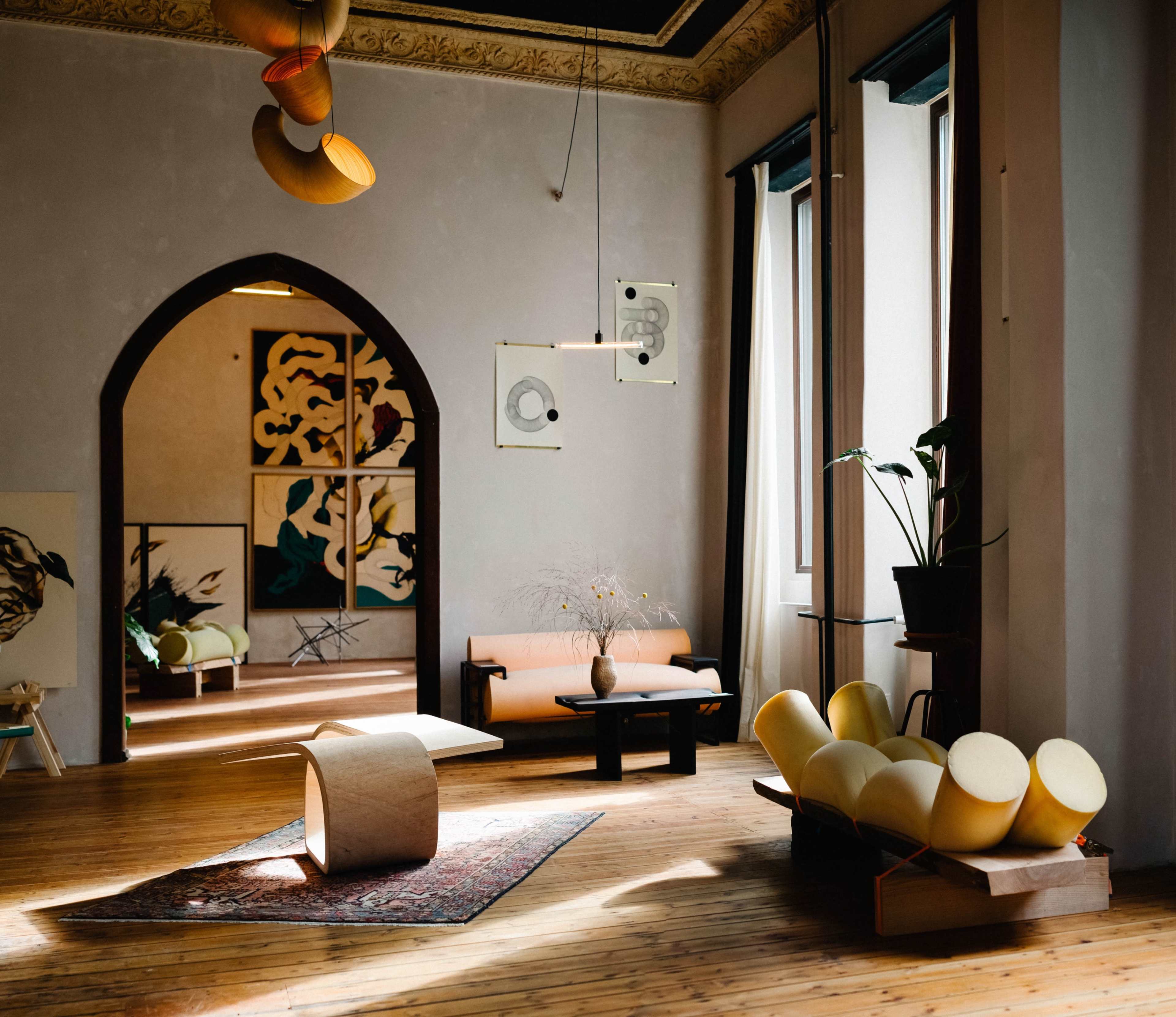 The image depicts a spacious room with wooden floors, large windows, and modern furniture, including a curved seating arrangement and abstract artwork on the walls.