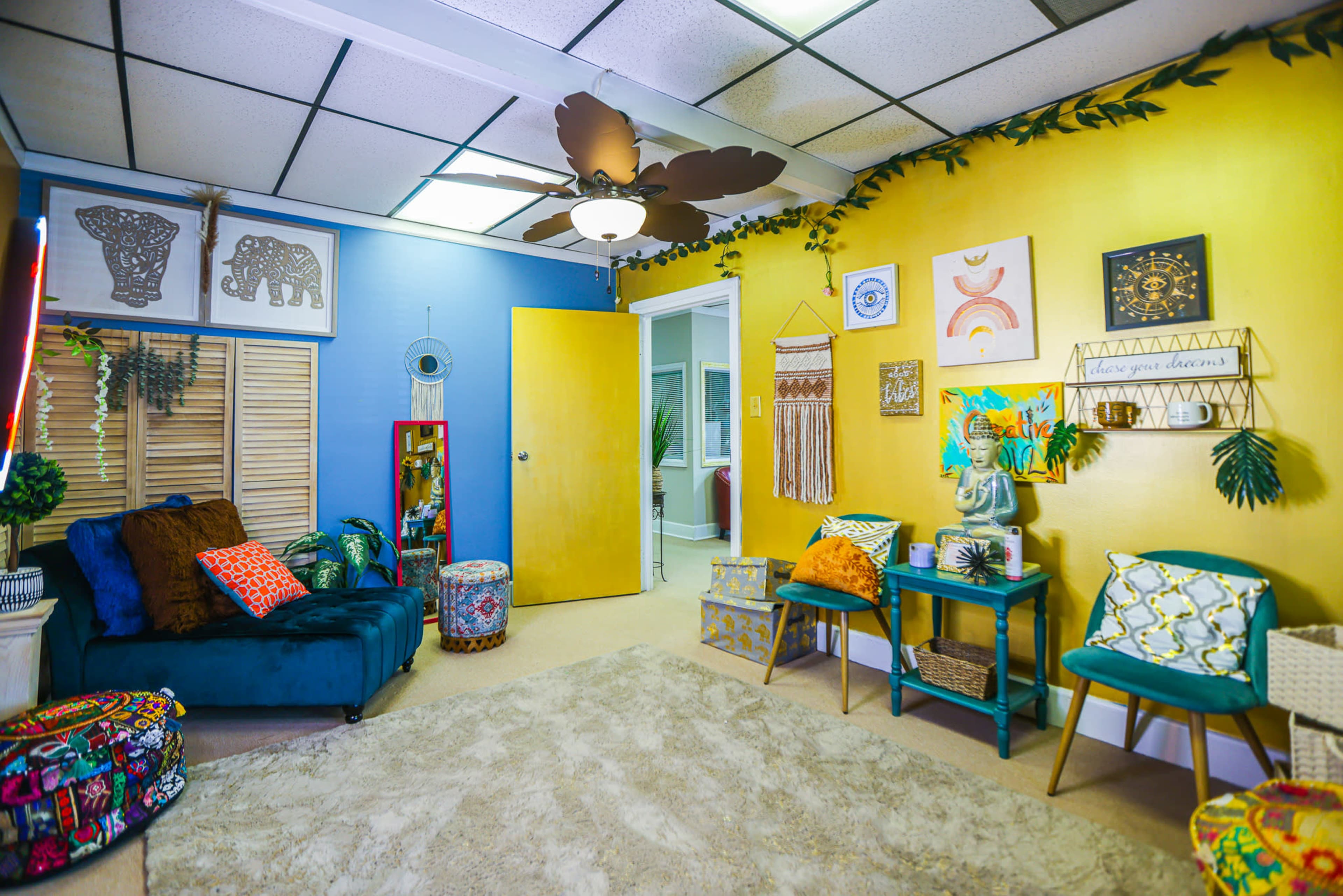 The image shows a colorful room with a blue and yellow color scheme, featuring a couch, decorative wall art, a rug, and various plants.