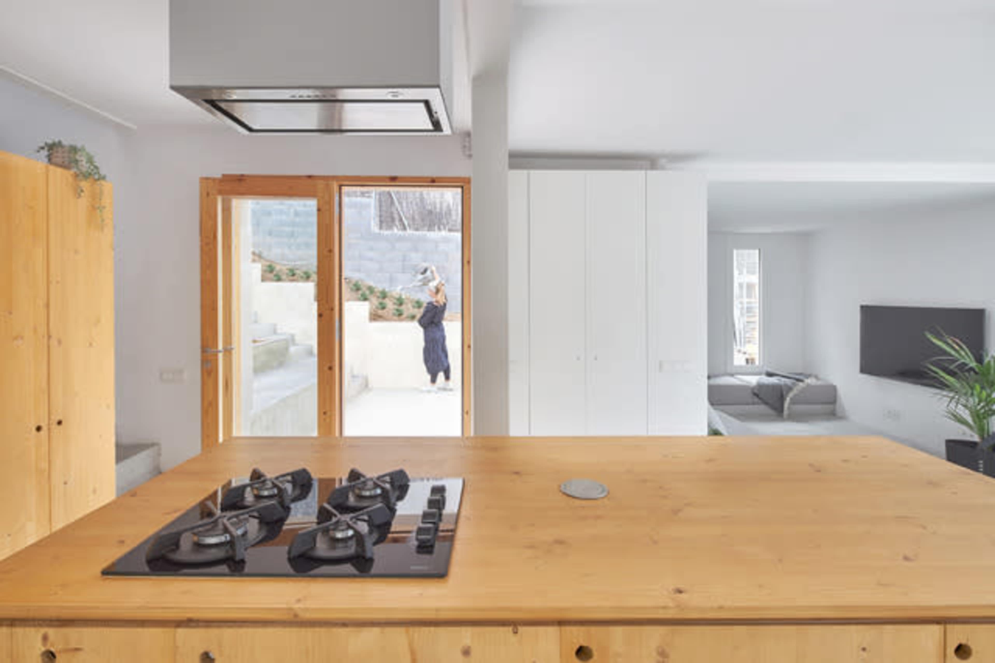 A kitchen features a wooden island with a gas stove, overlooking a living area with a television and a visible outdoor space through glass doors.