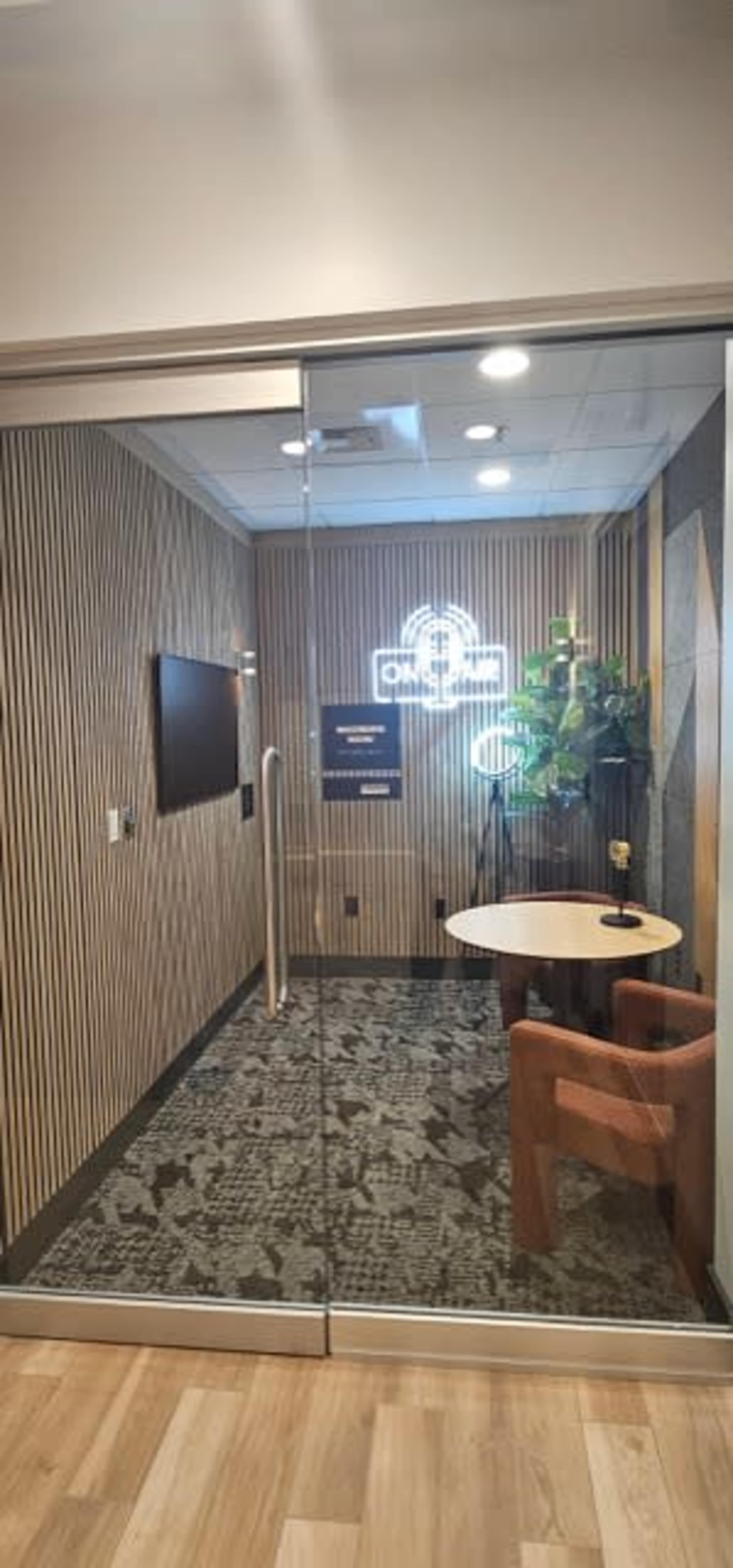 A glass-walled office features a small round table, two chairs, a wall-mounted screen, and a decorative plant against a wooden panel backdrop.