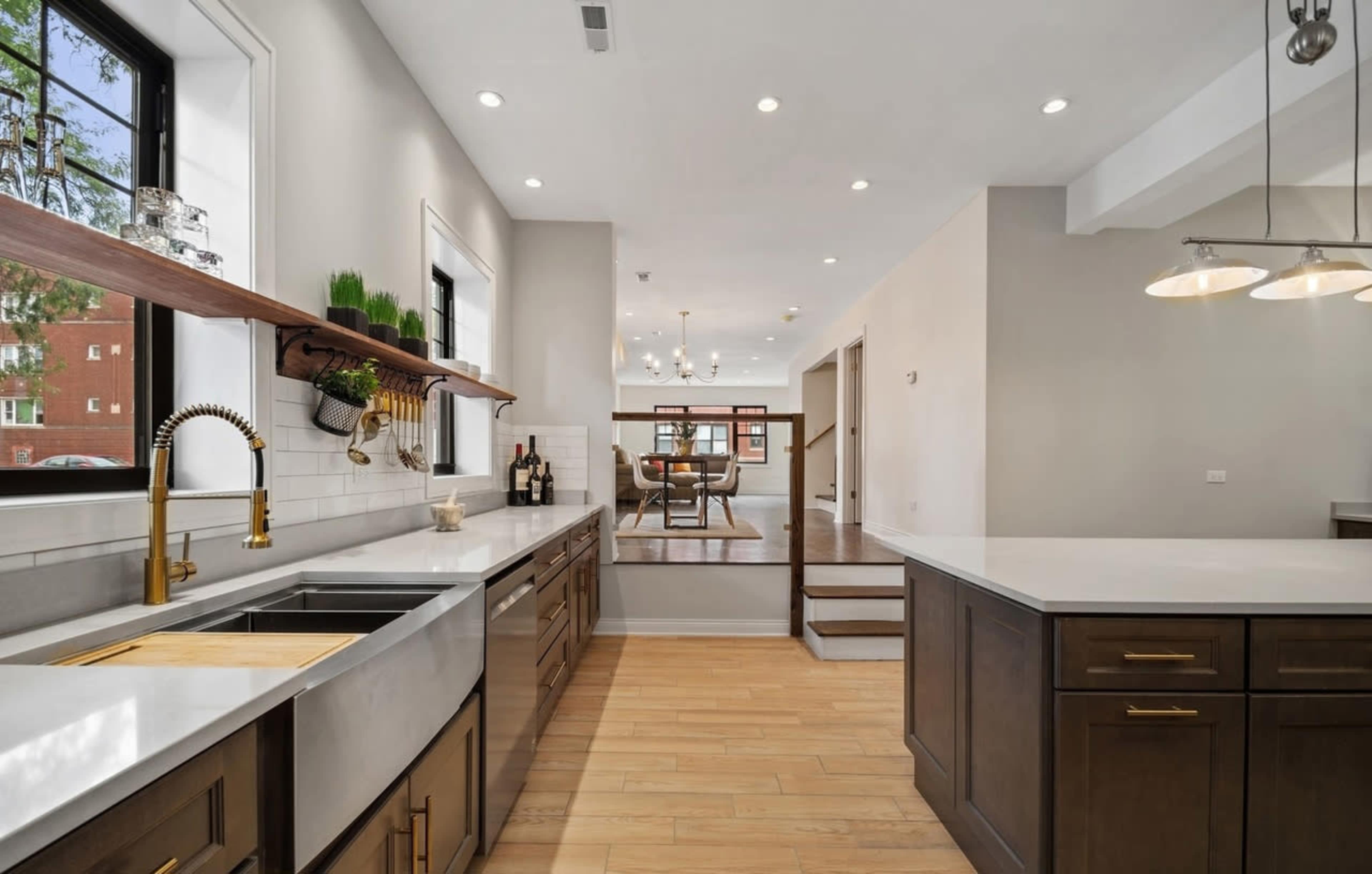 The image shows a modern kitchen with a large island, stainless steel fixtures, and wooden shelves containing decorative items.