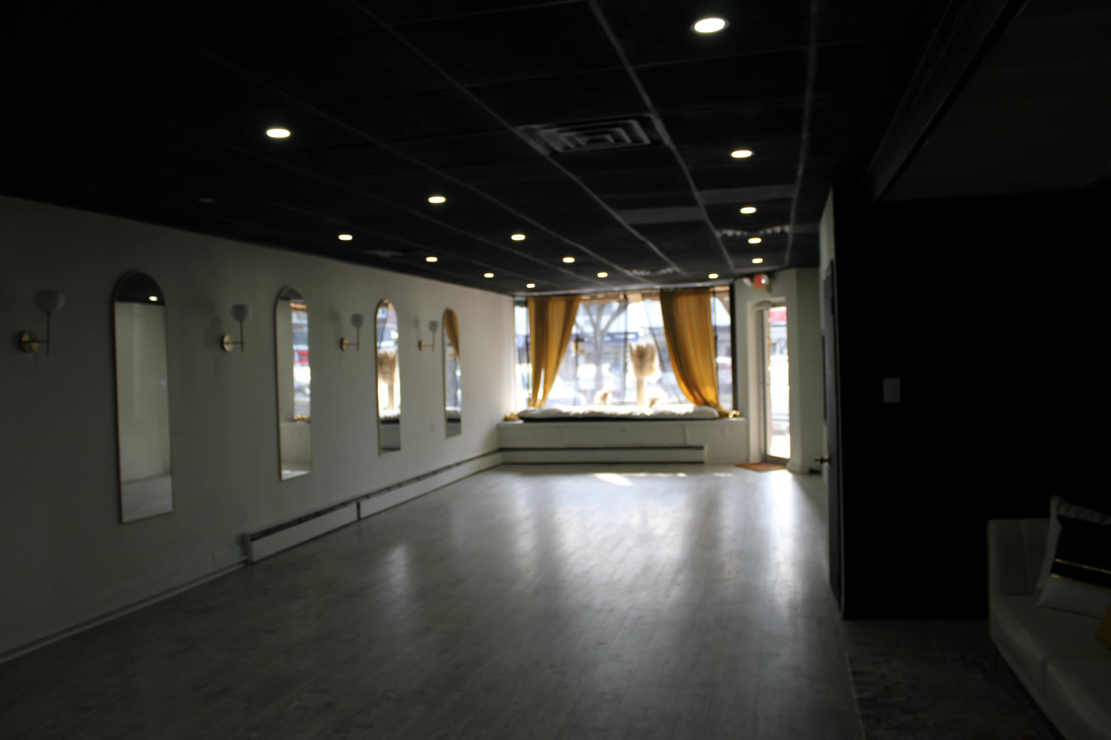 The image shows an empty room with large windows and mirrors on the walls, illuminated by overhead lights.