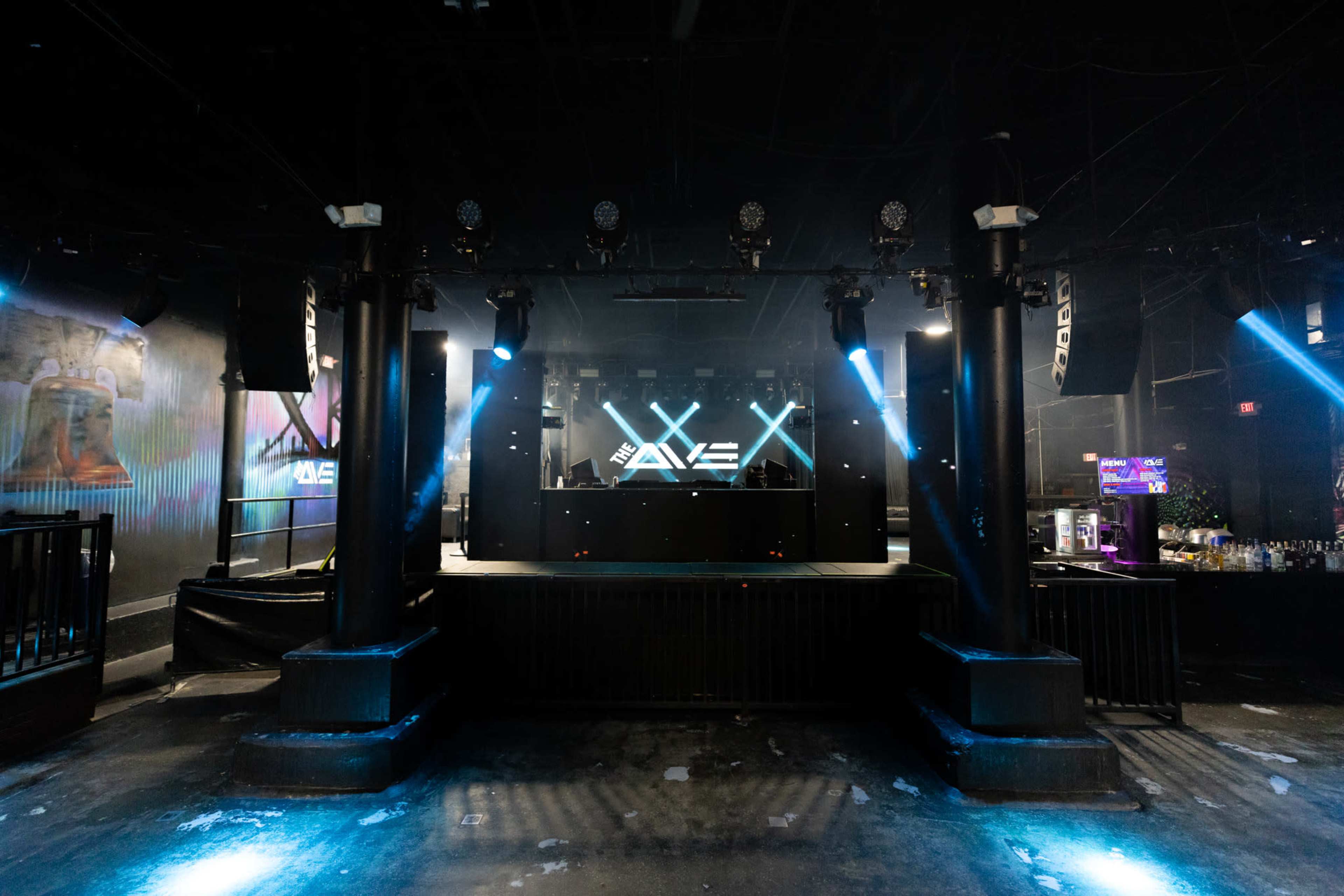 The image shows an empty nightclub interior with a DJ booth at the front, surrounded by large speakers and illuminated by stage lights.