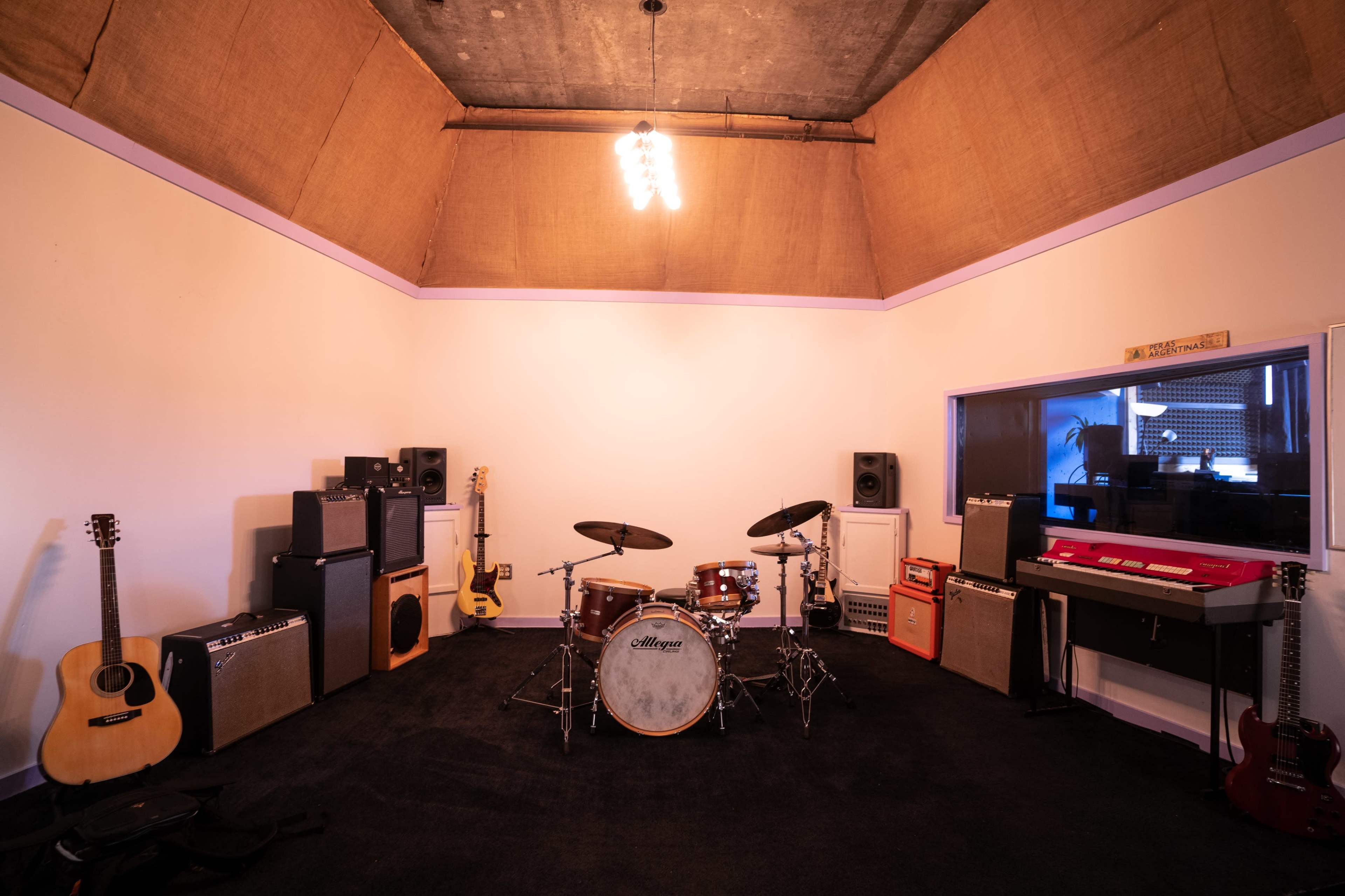 A music rehearsal studio featuring a drum set, amplifiers, guitars, a keyboard, and soundproofing elements on the walls.
