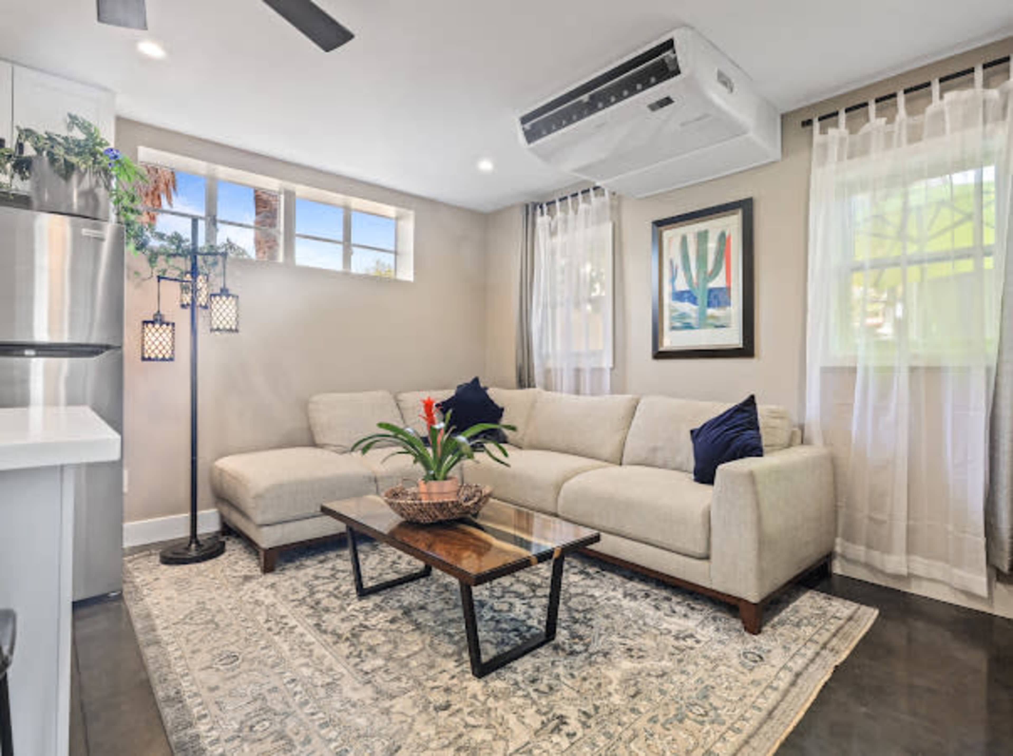 A modern living room features a beige sectional sofa, a wooden coffee table, and a window with sheer curtains, complemented by indoor plants and decorative lighting.