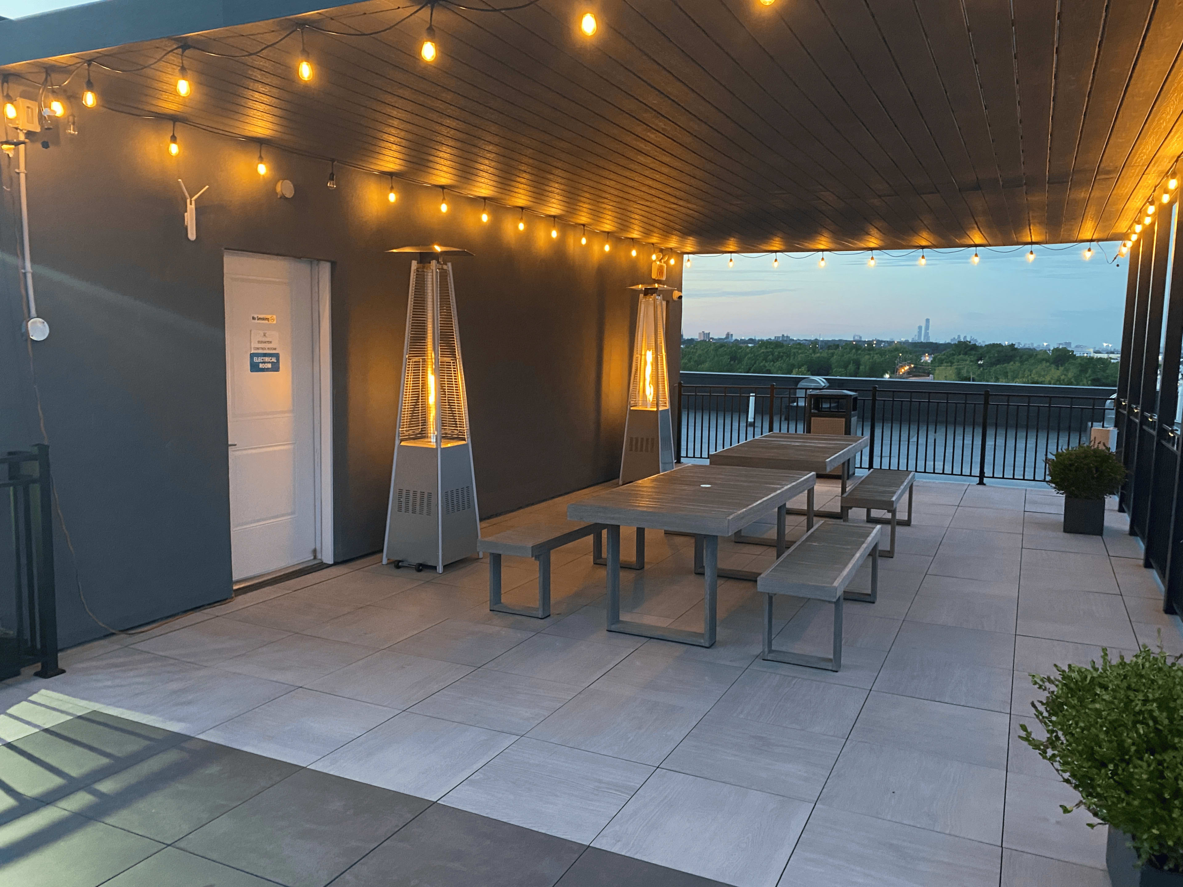 A rooftop terrace with string lights, two outdoor heaters, and a long table with benches overlooking a body of water.