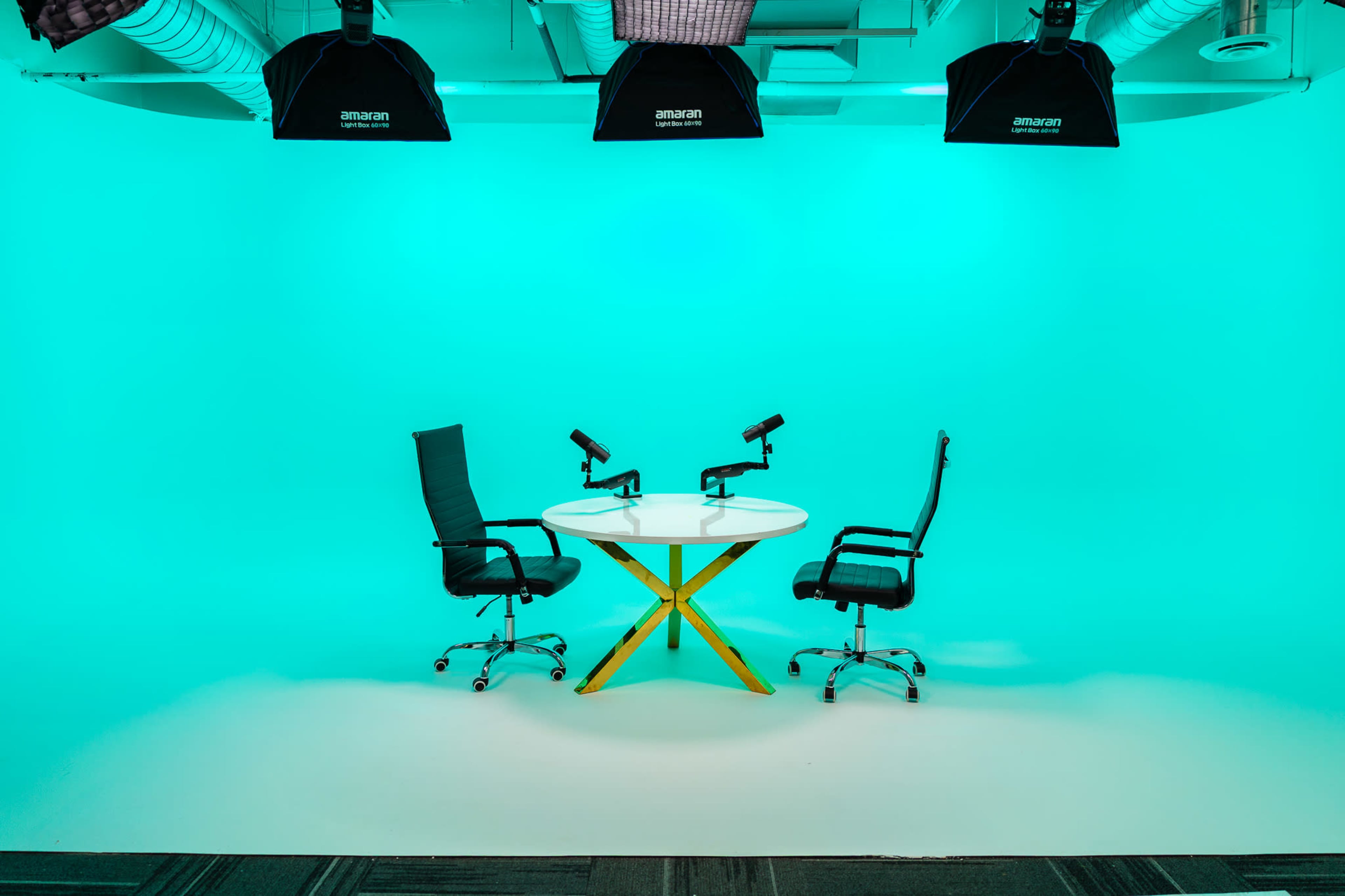 The image shows a brightly lit studio setup featuring a round table with two chairs and microphones positioned on each side, set against a teal backdrop.