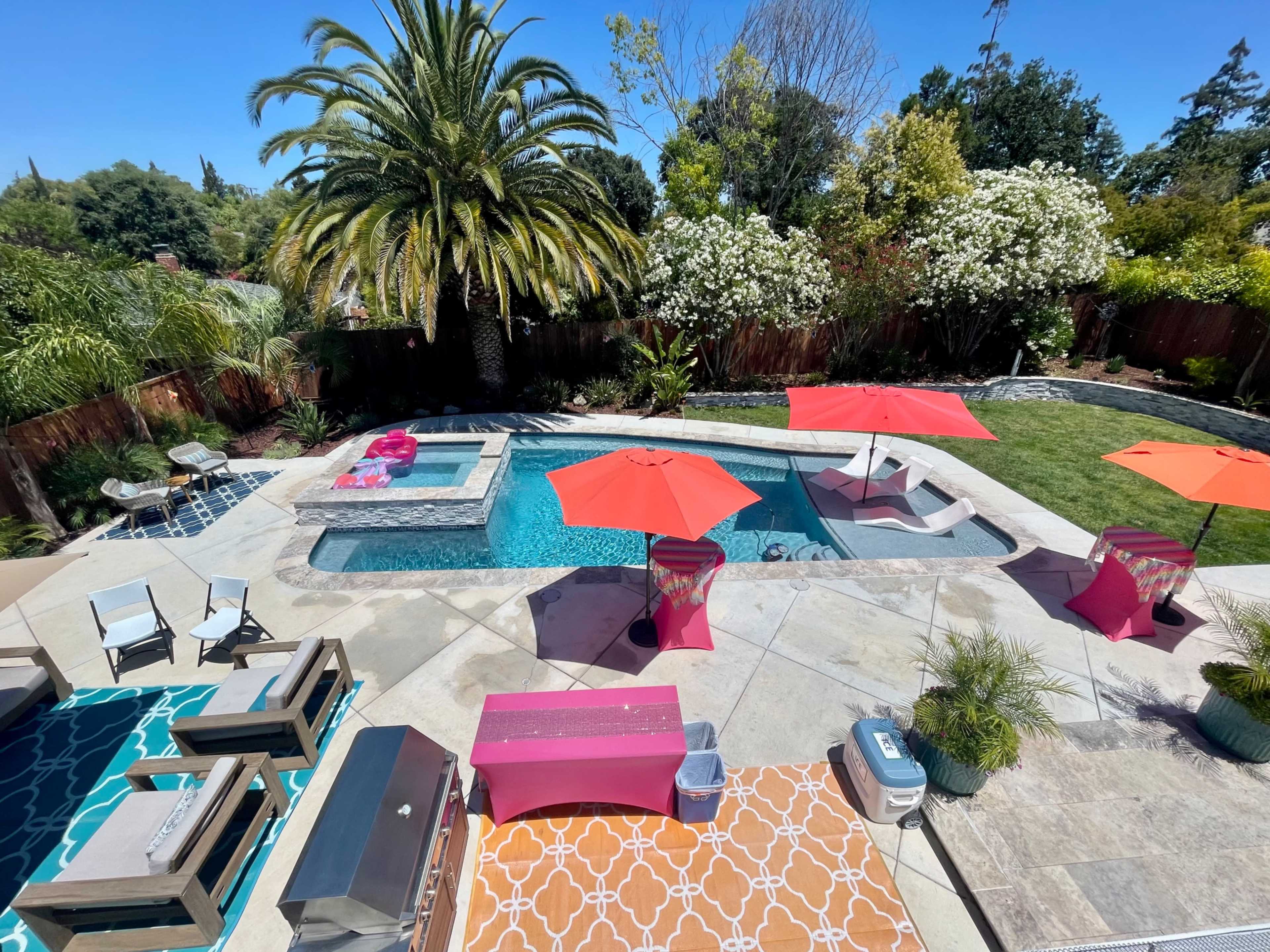 The image shows a backyard with a swimming pool, surrounded by lounge chairs, umbrellas, and palm trees.