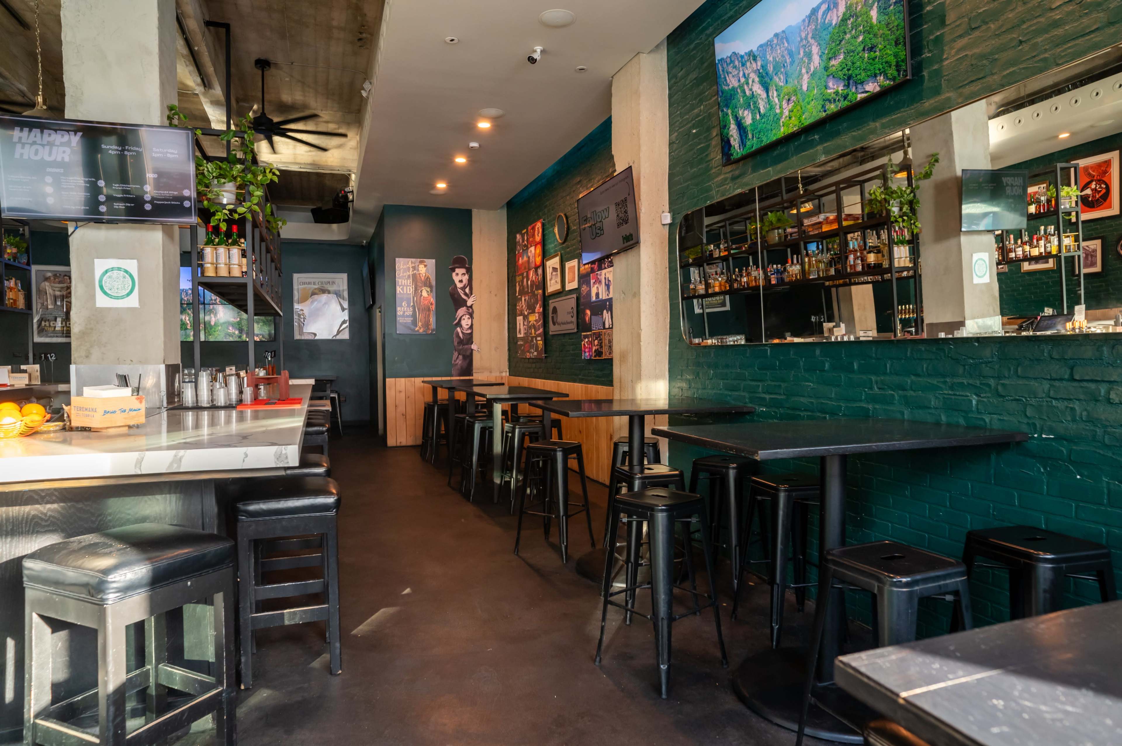 The image shows a modern bar and dining area with green walls, high tables, and a mirrored back wall displaying various drinks and decor.