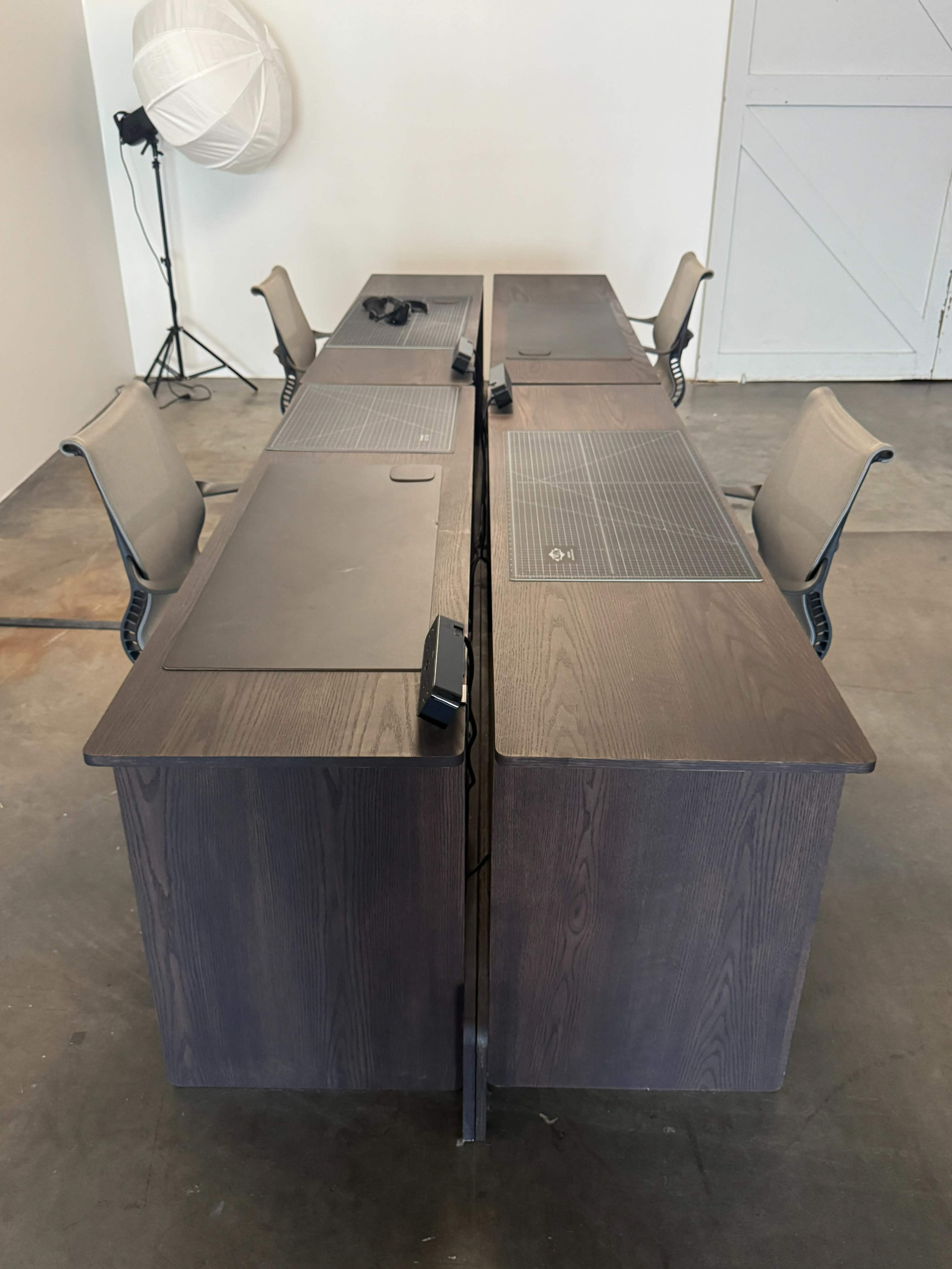 The image shows two large desks arranged in an L-shape, with four ergonomic chairs positioned around them.