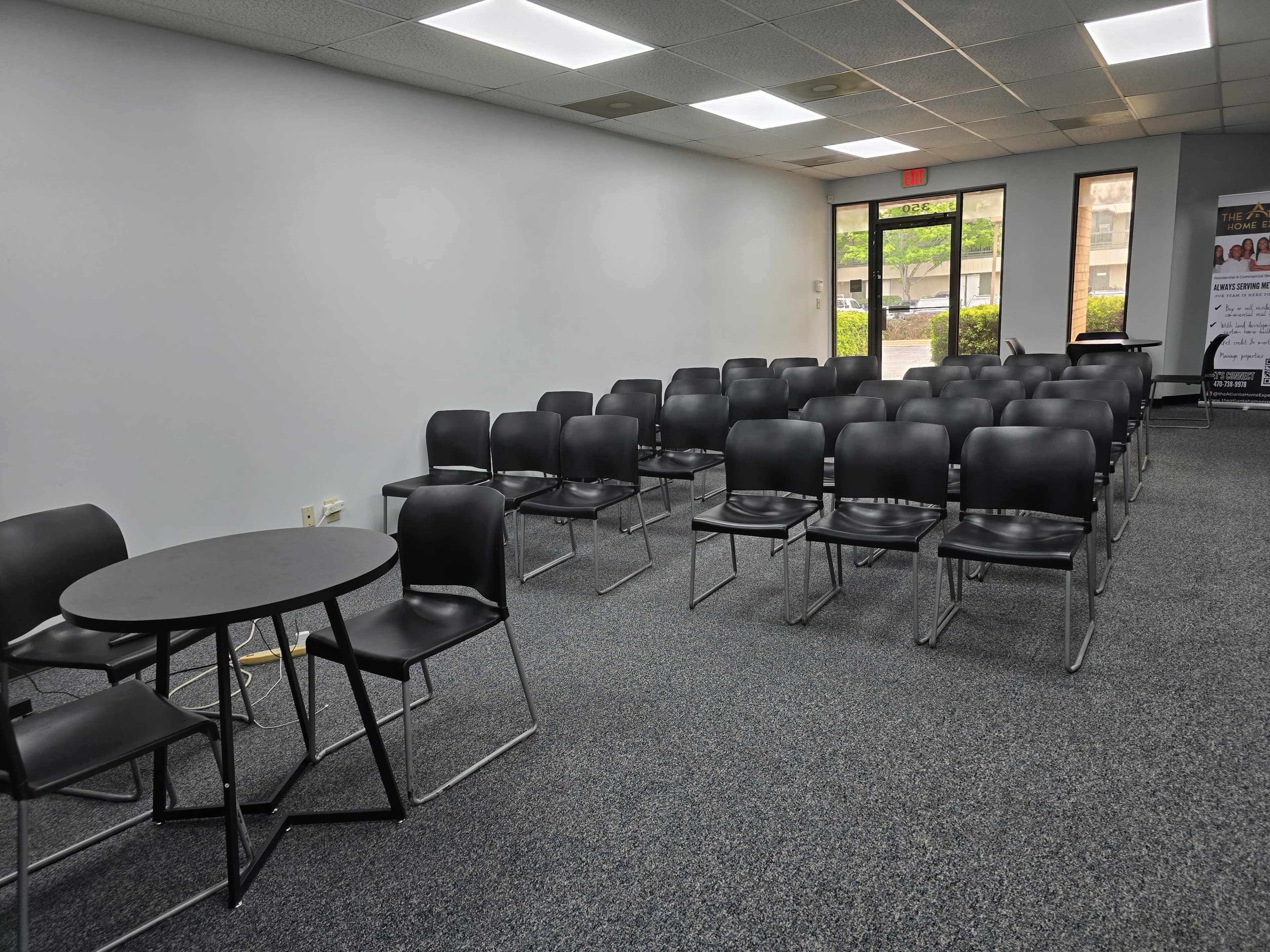 The image shows a spacious room with a row of black chairs arranged in several rows, accompanied by a small round table positioned on the left side.