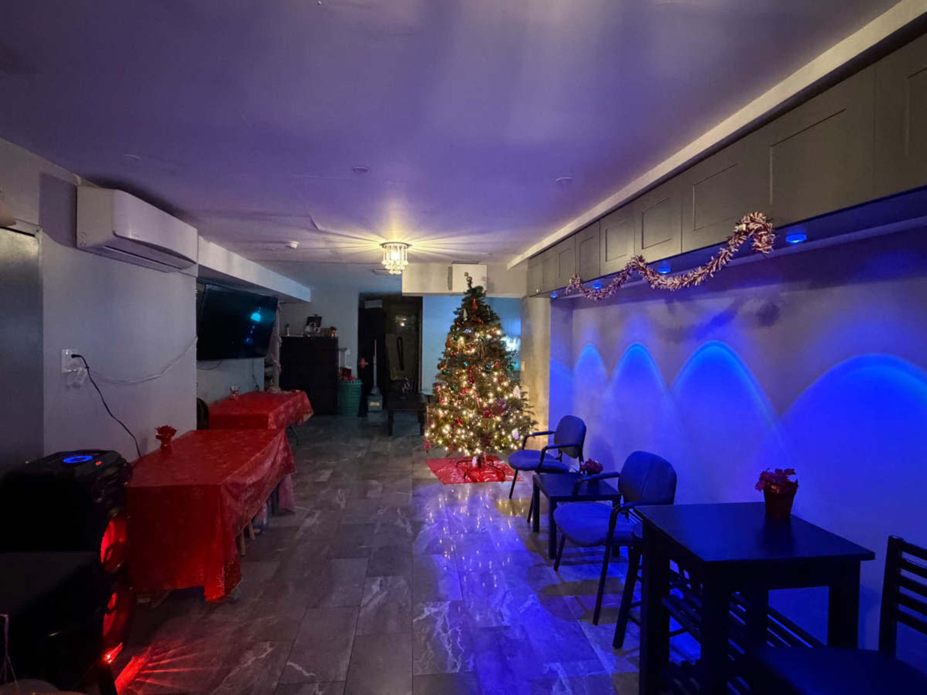 The image shows a festive indoor space decorated with a Christmas tree, dining tables covered in red cloth, and blue lighting along the walls.