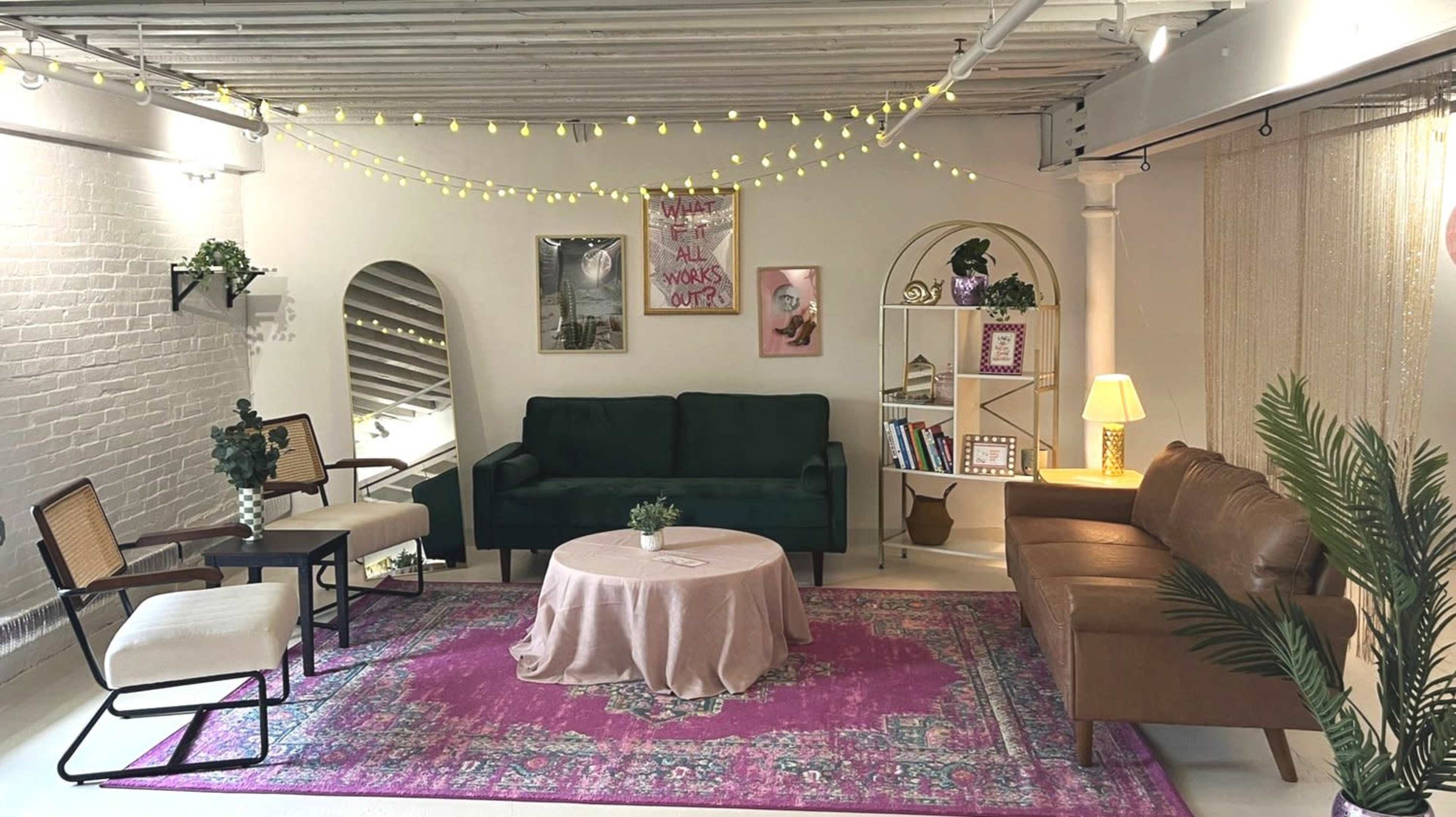 A cozy lounge area featuring green and brown sofas, a circular coffee table, a pink rug, and decorative shelves against a light-colored wall adorned with string lights.
